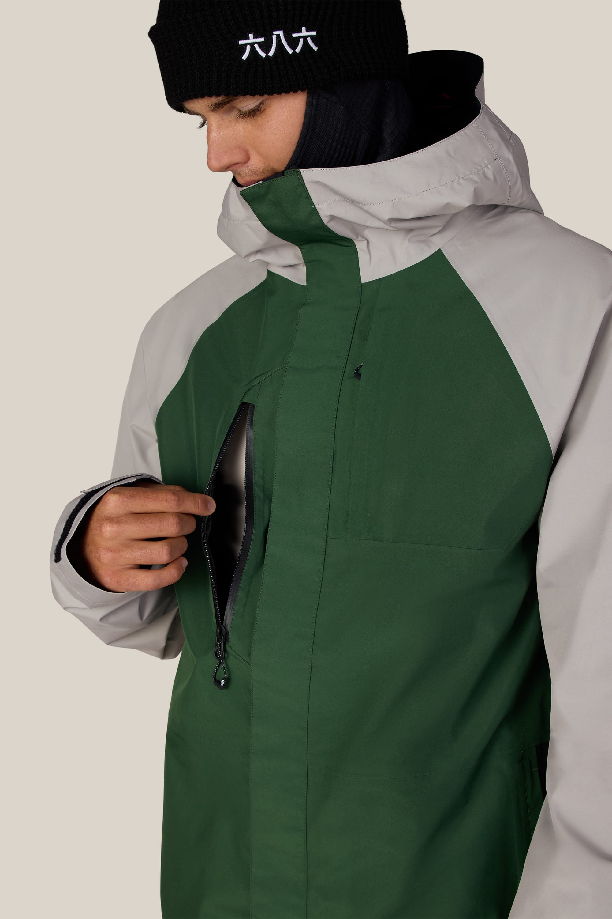 A person wearing the 686 Men’s GORE-TEX® Core Shell Jacket in a two-tone green and gray colorway unzips the chest pocket. They are also wearing a black beanie with white characters and looking down at the pocket.