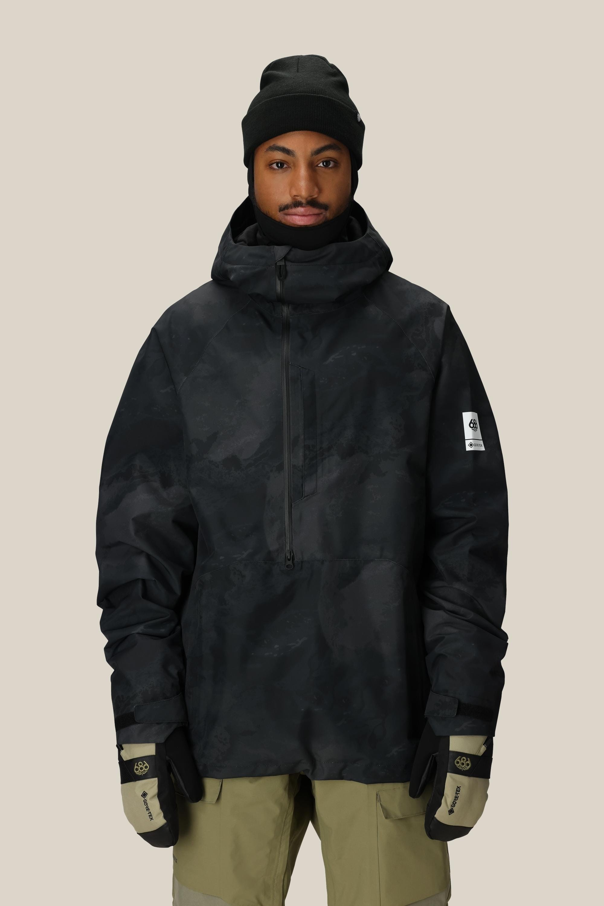 A person wearing the 686 Mens GORE-TEX Fragment Shell Anorak, black beanie, beige pants, and black-and-beige gloves stands against a plain light background.