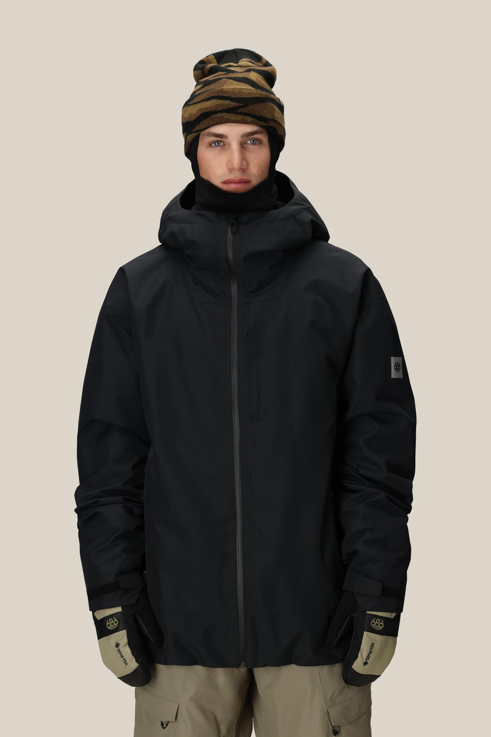 A person wearing a 686 Mens Gateway Shell Jacket in black with a hood, a patterned beanie, beige gloves, and beige pants stands facing forward against a plain light background.