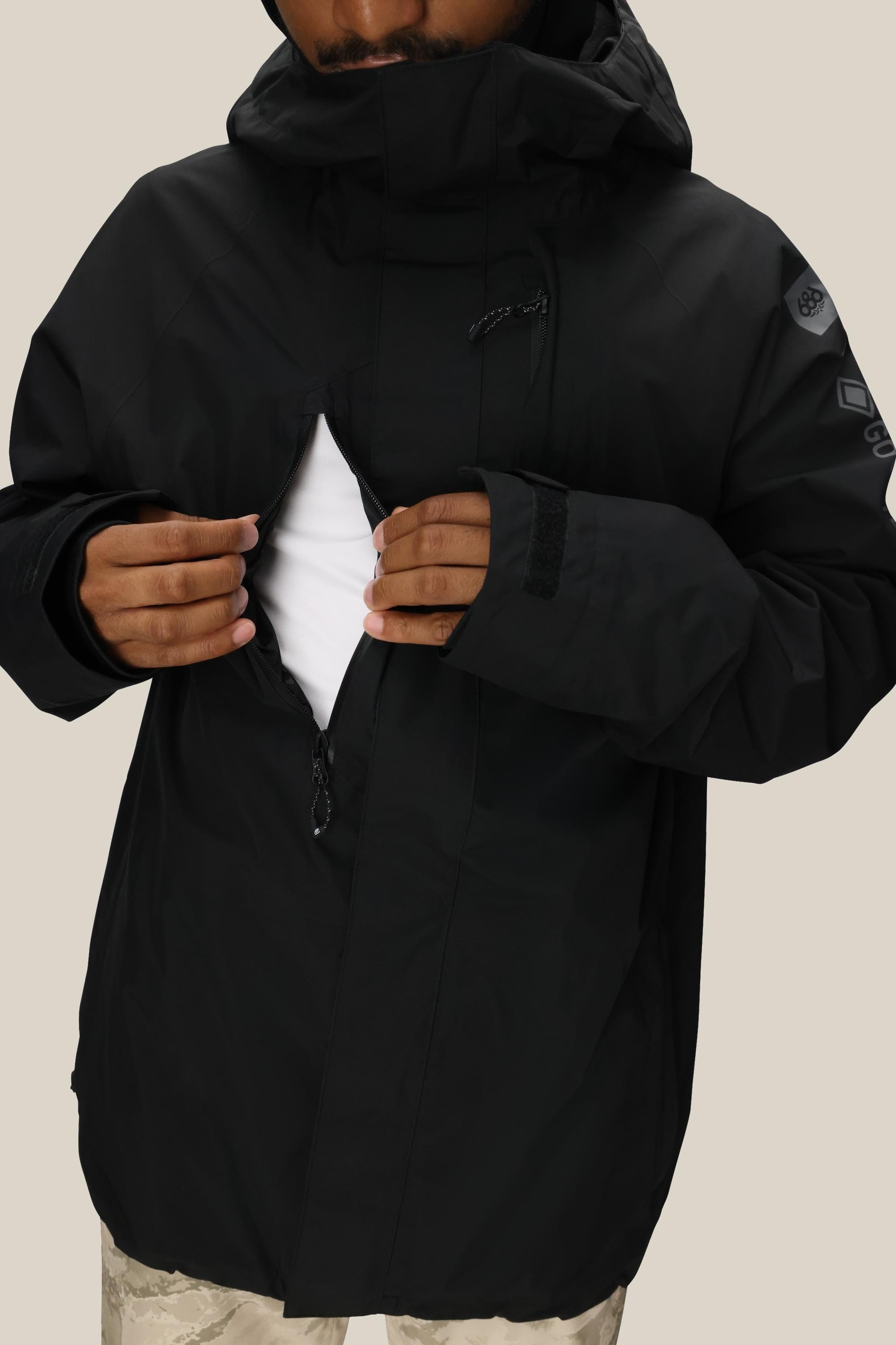 A person wearing the 686 Men’s GORE-TEX® Core Insulated Jacket in black partially unzips the front to show a white shirt underneath. Only the lower part of their face is visible, set against a plain beige background.
