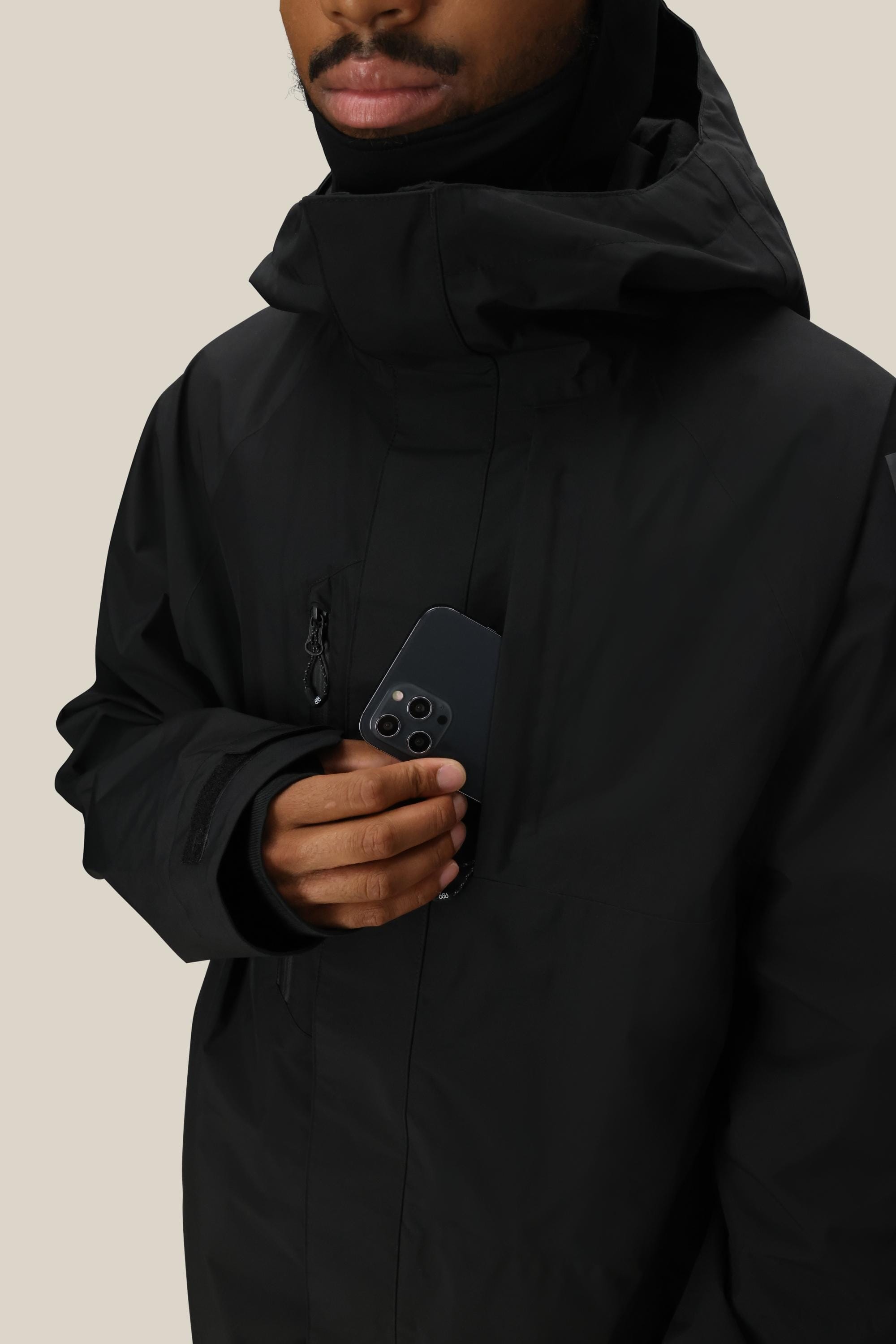 A person wearing the 686 Men’s GORE-TEX® Core Insulated Jacket in black slips a black smartphone into the jacket’s chest pocket. The background is plain and light-colored, with only the lower half of the person’s face visible.