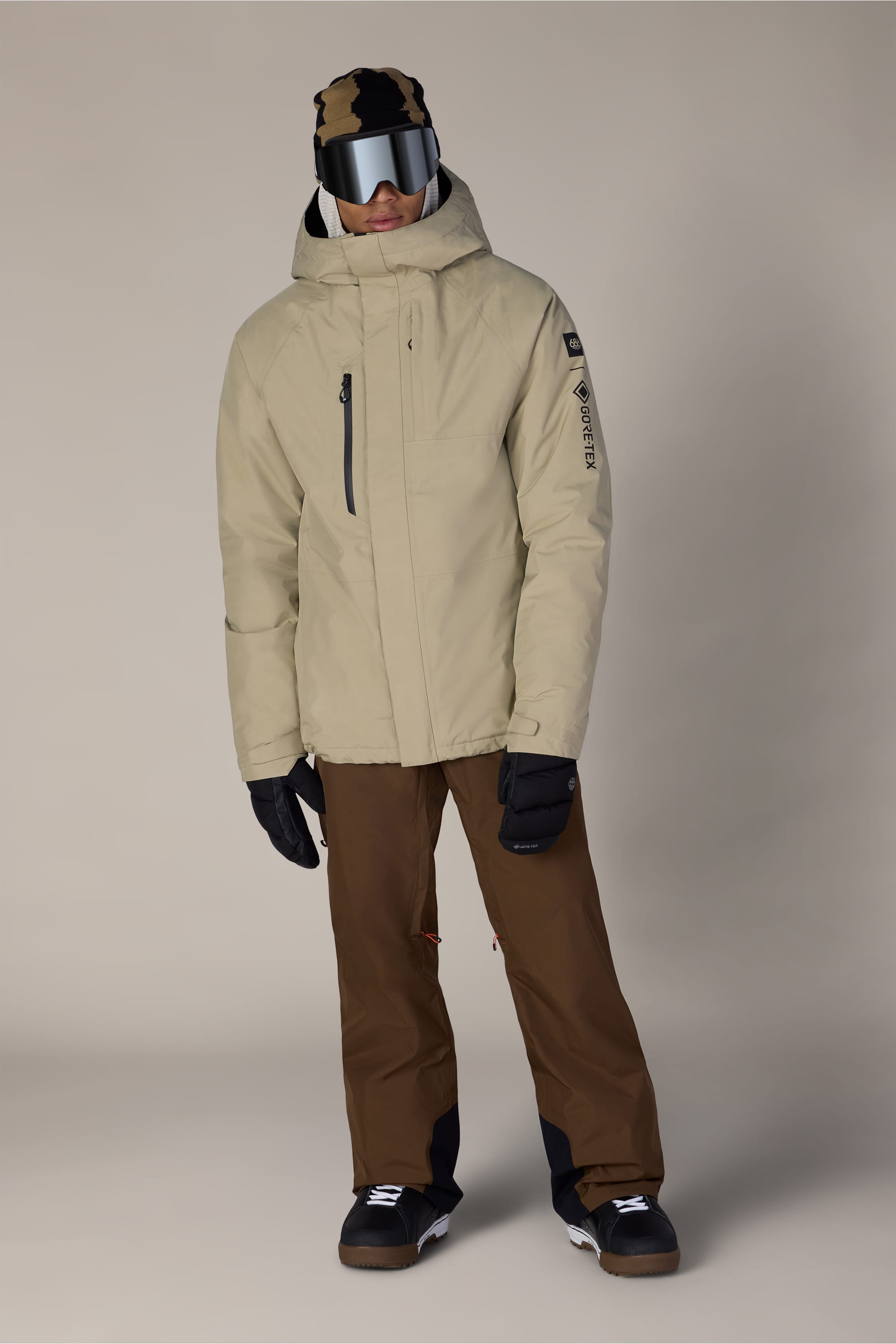 A person wearing the 686 Men’s GORE-TEX® Core Insulated Jacket in beige, paired with brown ski pants, black gloves and boots, a tan and black beanie, and large ski goggles, standing against a plain light background.