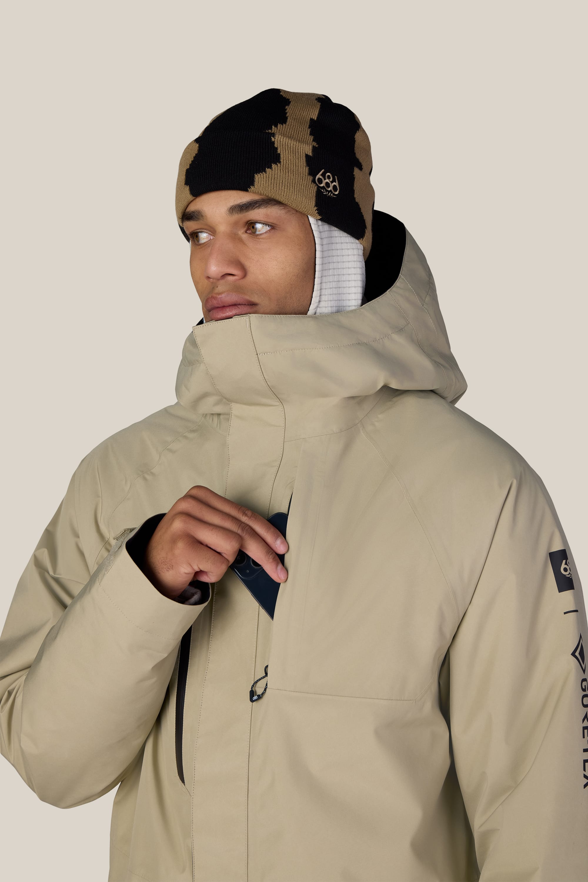 A person wearing the 686 Men’s GORE-TEX® Core Insulated Jacket in tan and a brown-and-black beanie looks to the side while zipping up the jacket. The background is plain and light-colored.