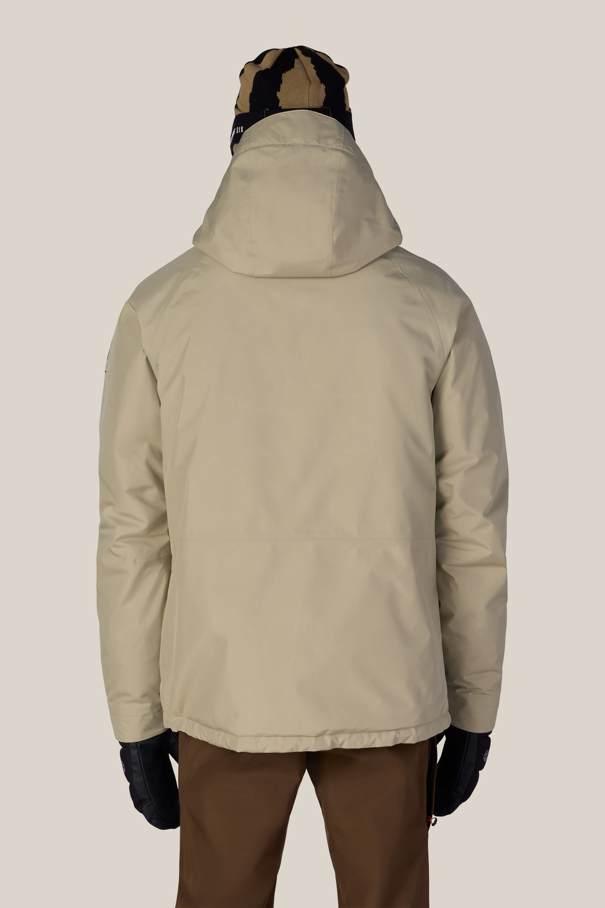 A person wearing the 686 Men’s GORE-TEX® Core Insulated Jacket in beige, paired with brown pants, black gloves, and a black-and-tan beanie, stands with their back to the camera against a plain light background.