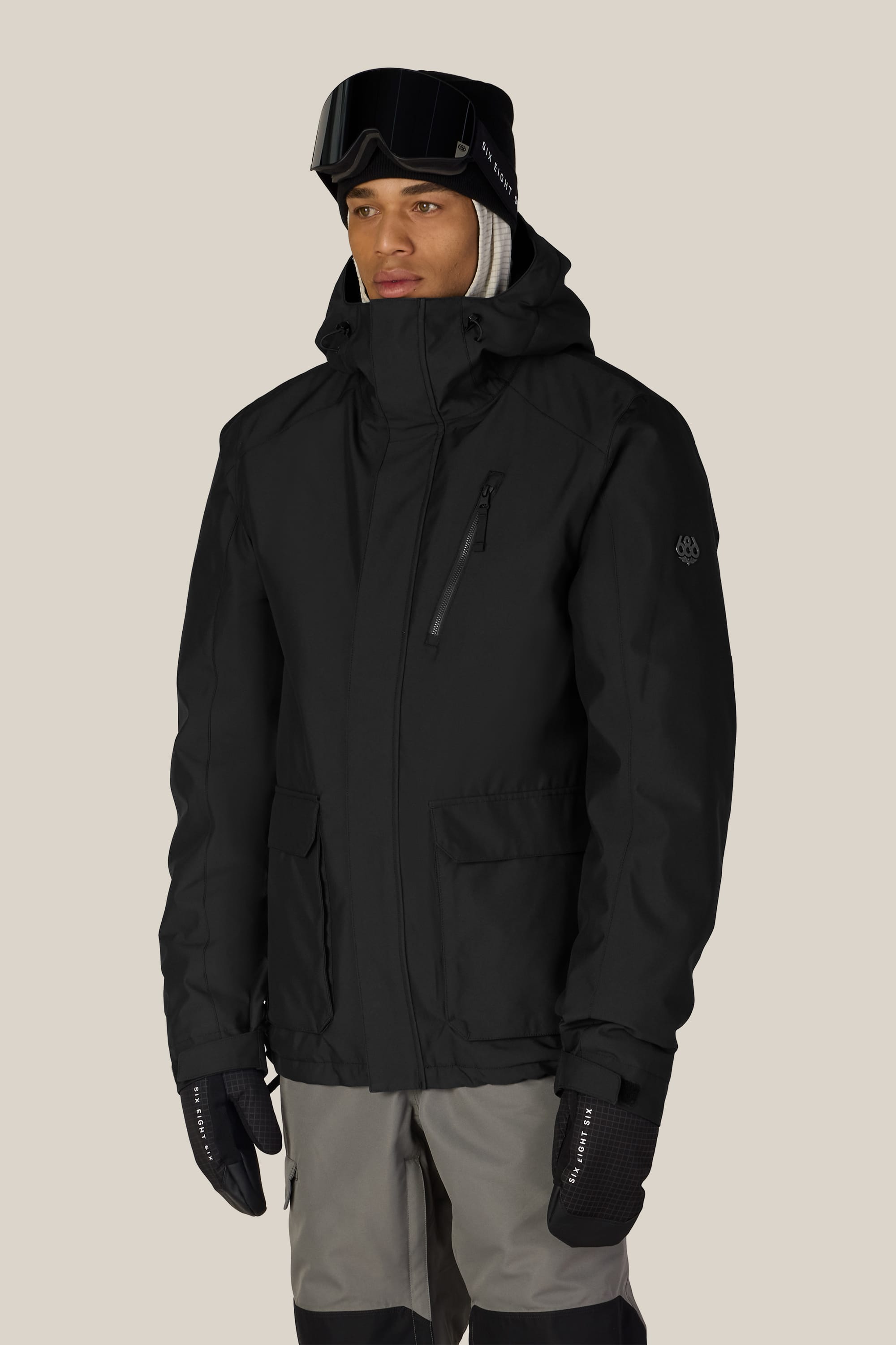 Wearing the 686 Men’s Nightline™ Insulated Jacket by 686, along with black gloves, gray and black snow pants, a black beanie, and ski goggles, a person stands against a plain light background.
