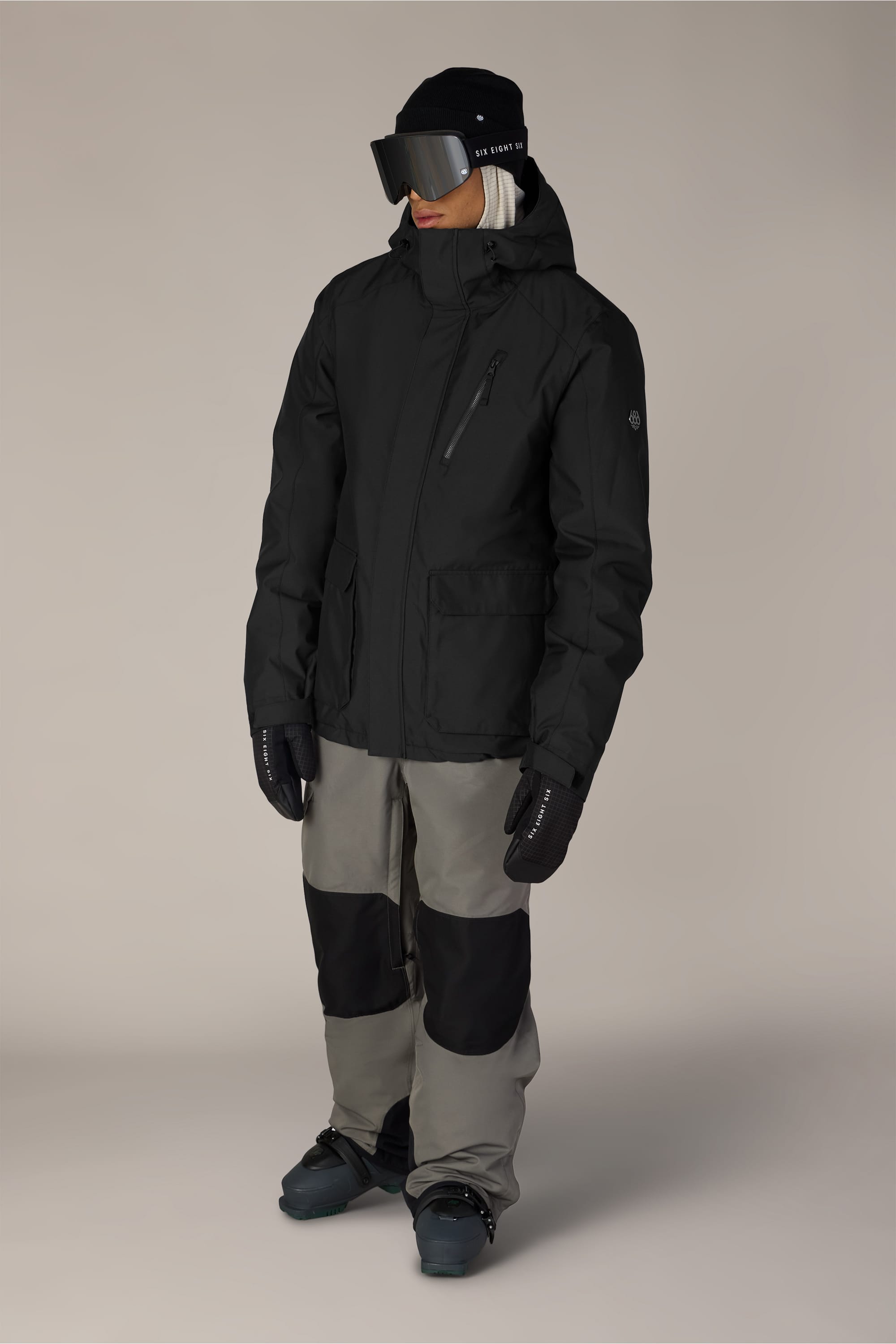 A person stands against a plain background wearing the 686 Men’s Nightline™ Insulated Jacket by 686, paired with gray pants featuring black knee patches, black gloves, dark ski goggles, and a black beanie.