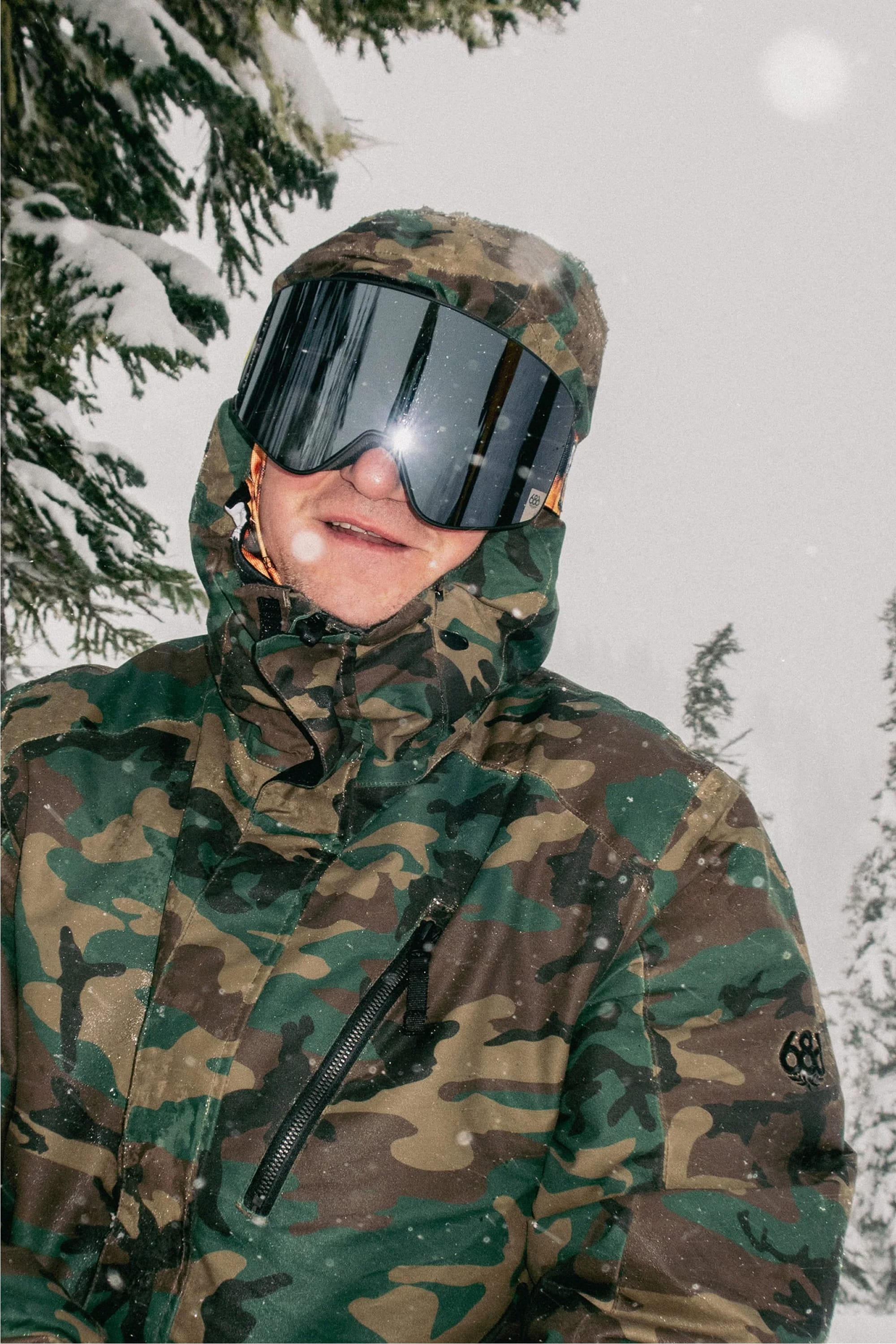A person wearing the 686 Men’s Nightline™ Insulated Jacket and reflective ski goggles stands in a snowy forest, surrounded by snow-covered trees and falling snowflakes.