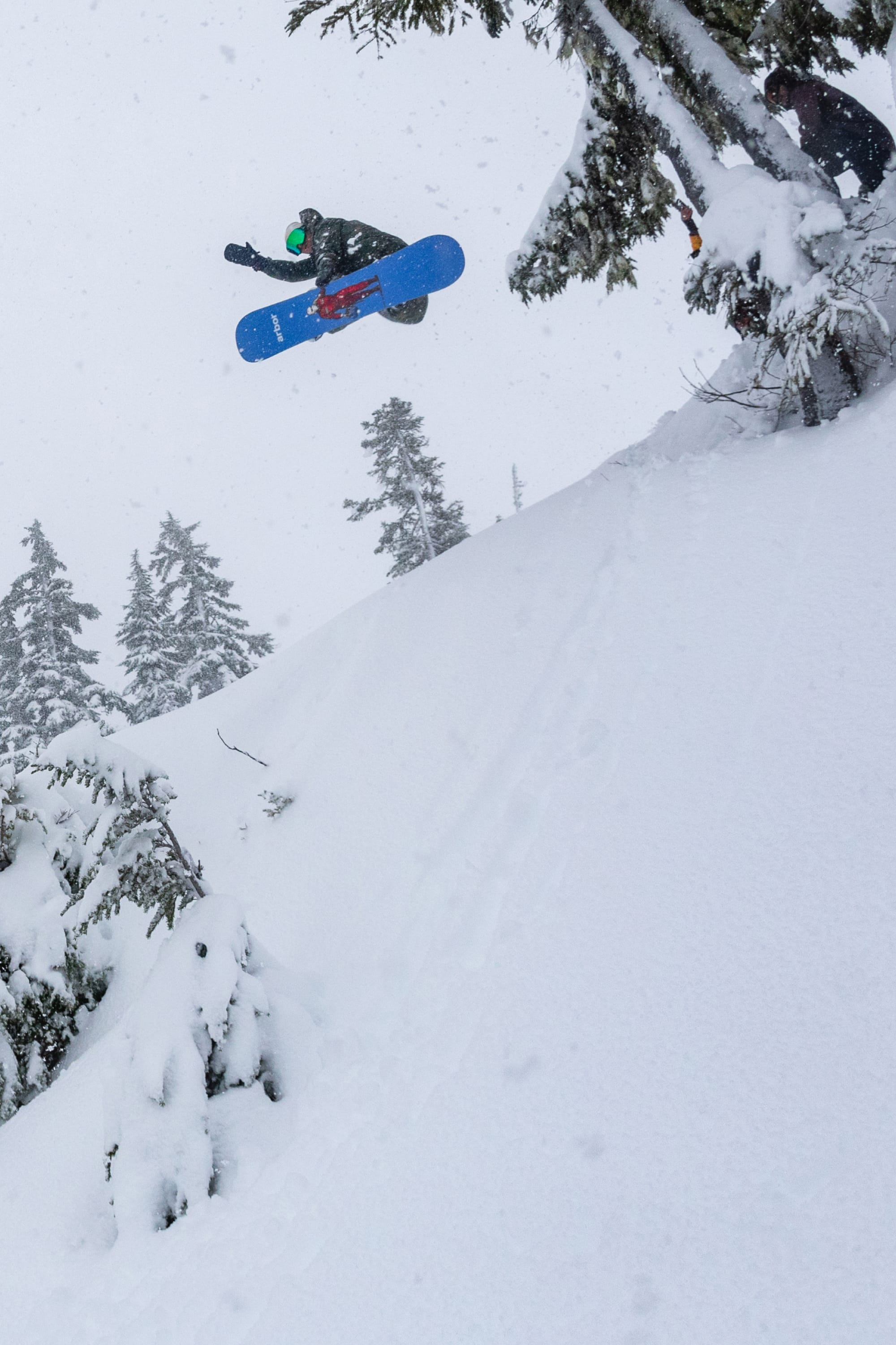 Wearing the 686 Men’s Nightline™ Insulated Jacket, a snowboarder launches off a snowy slope and soars through the air amid snow-covered trees on a cloudy winter day.