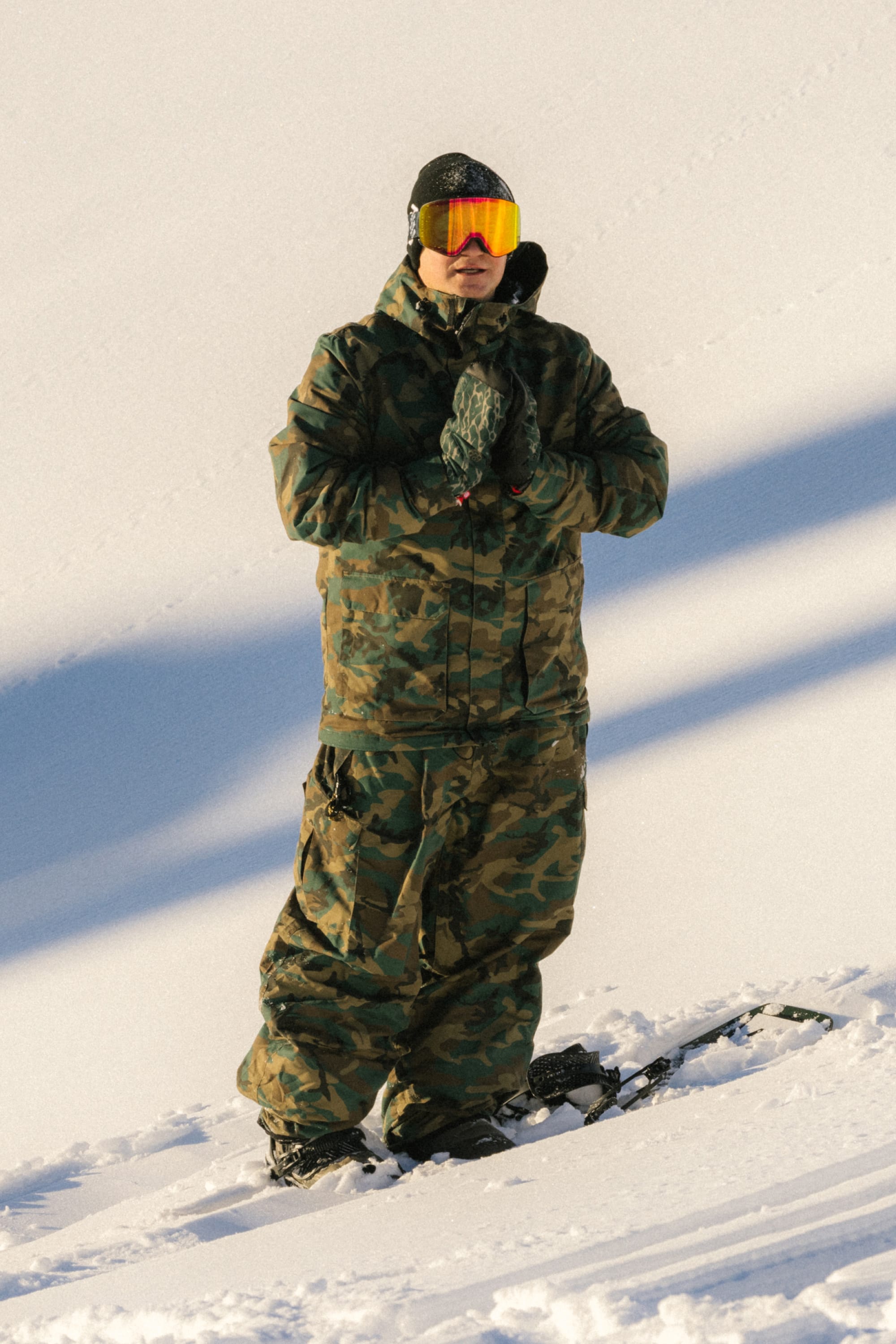 Clad in the 686 Men’s Nightline™ Insulated Jacket and reflective goggles, a person stands on snowy terrain with hands clasped beside a snowboard, shadows stretching across the winter landscape.