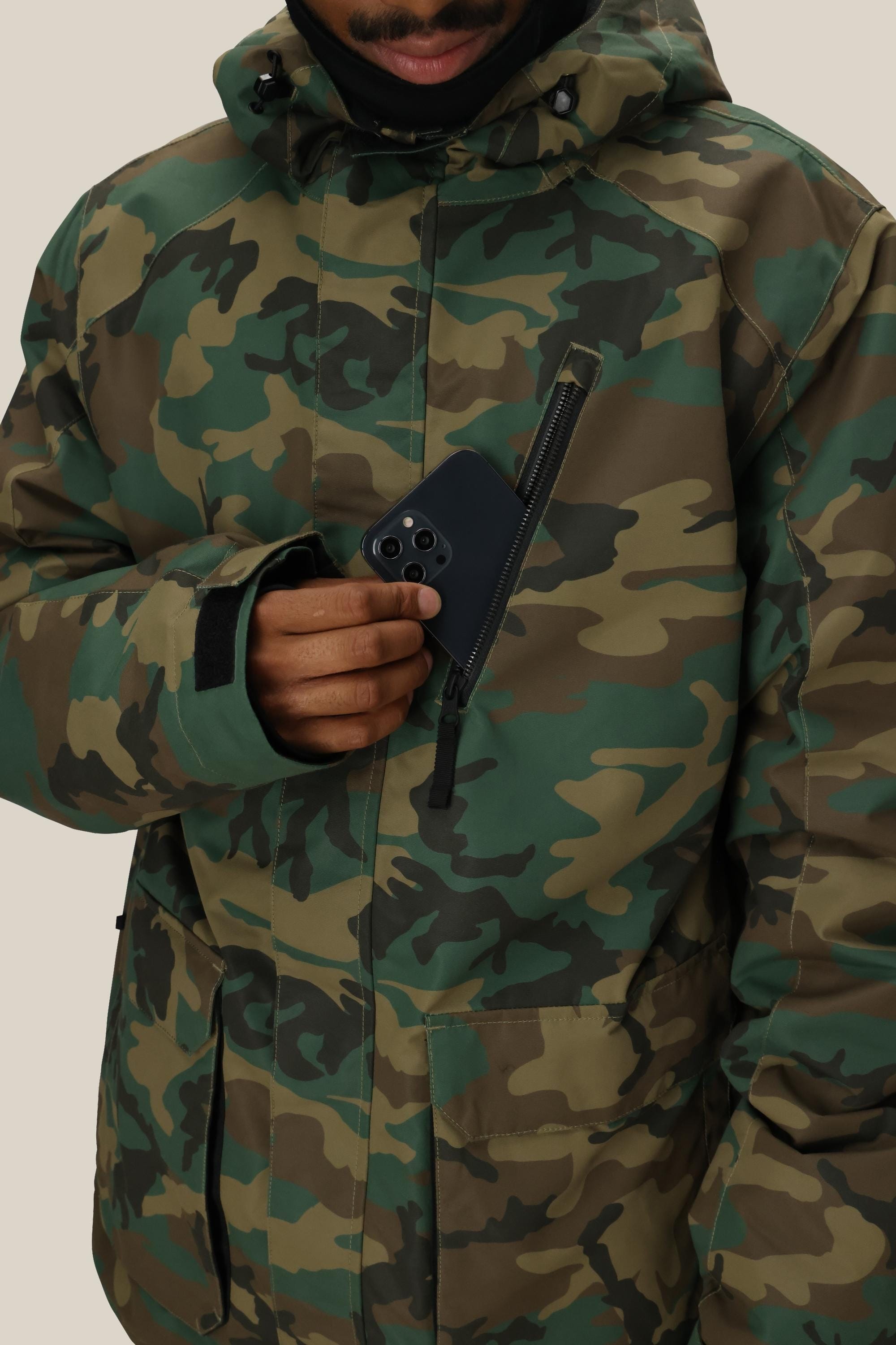 A person wearing the 686 Men’s Nightline™ Insulated Jacket in green camouflage places a black smartphone into the jacket’s zippered chest pocket. Their face is partially visible, set against a plain beige background.