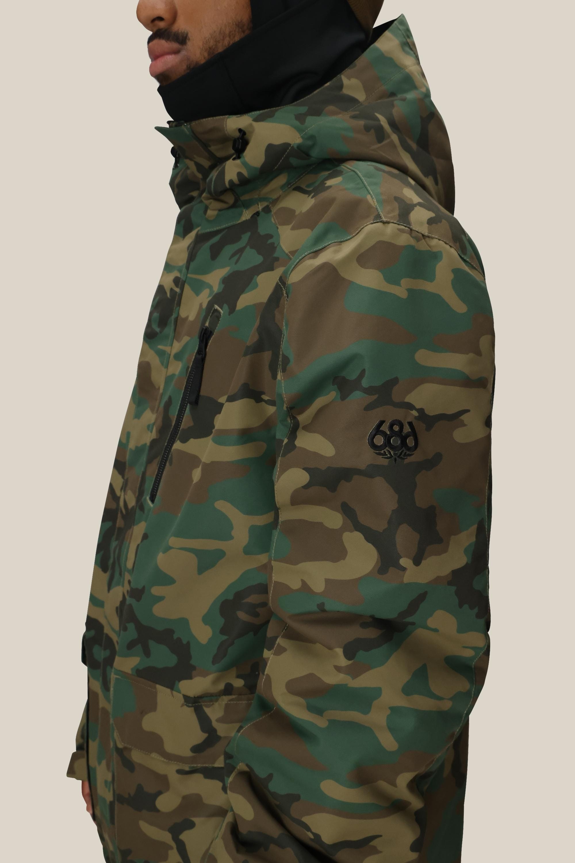 A person is wearing the 686 Men’s Nightline™ Insulated Jacket, a green and brown camouflage hooded jacket from 686, featuring a high collar and a black logo on the sleeve. The person’s face is mostly out of view.