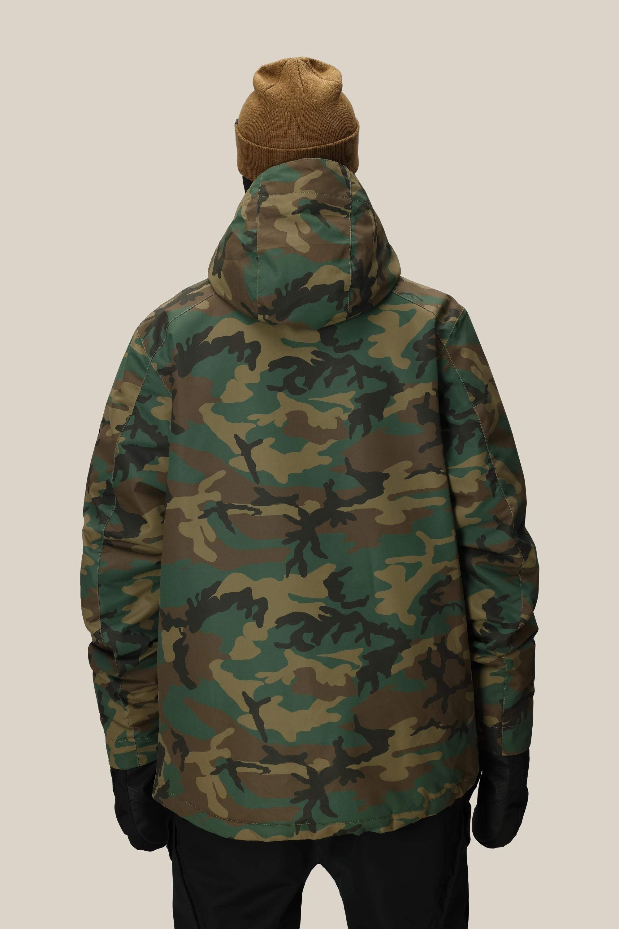 A person is seen from behind, wearing a brown beanie, black gloves, and the 686 Men’s Nightline™ Insulated Jacket with a hood in green camouflage by 686, set against a plain beige background.