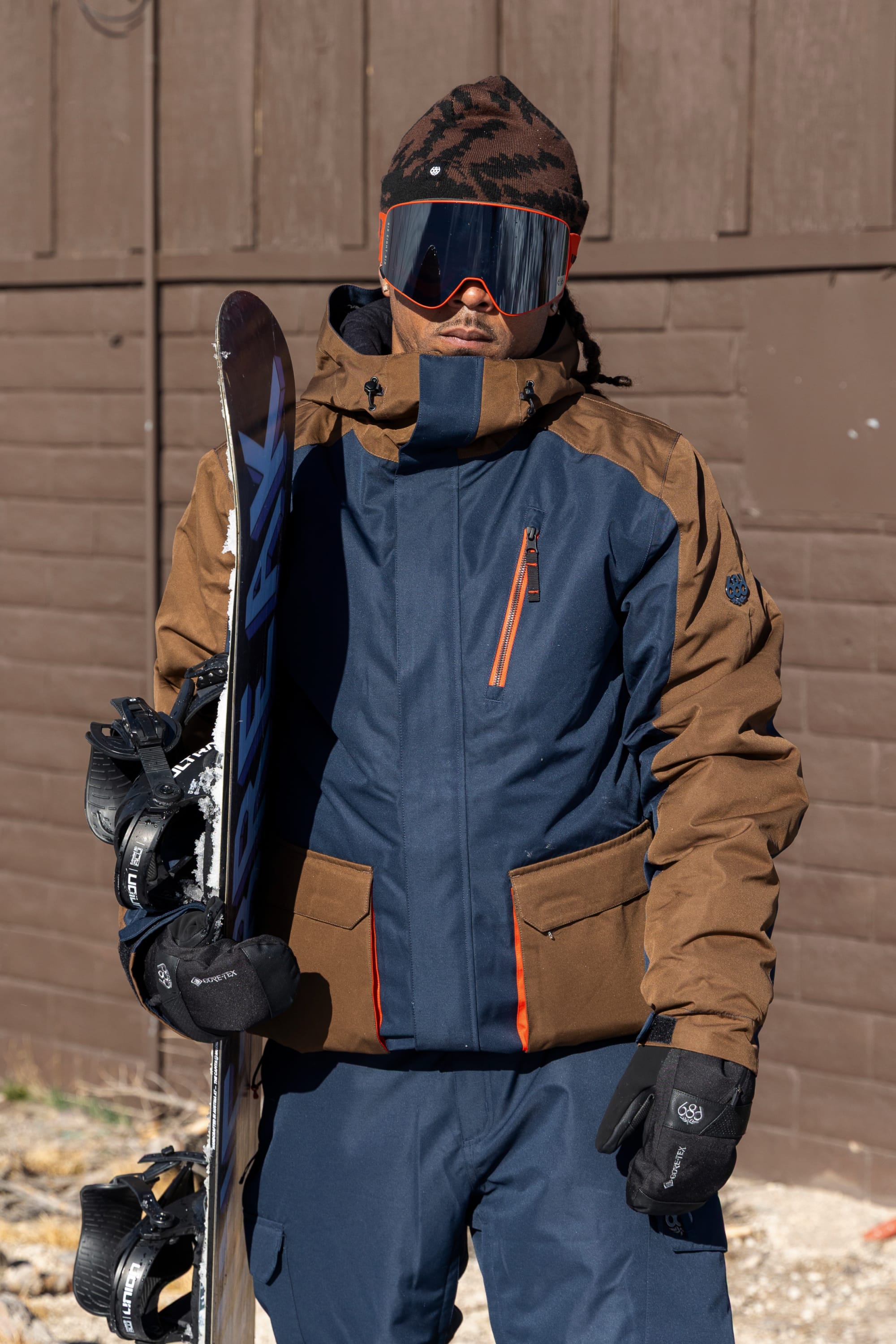A person wearing the 686 Men’s Nightline™ Insulated Jacket, along with gloves, goggles, and a beanie, stands outside in front of a brown wooden wall holding a snowboard.