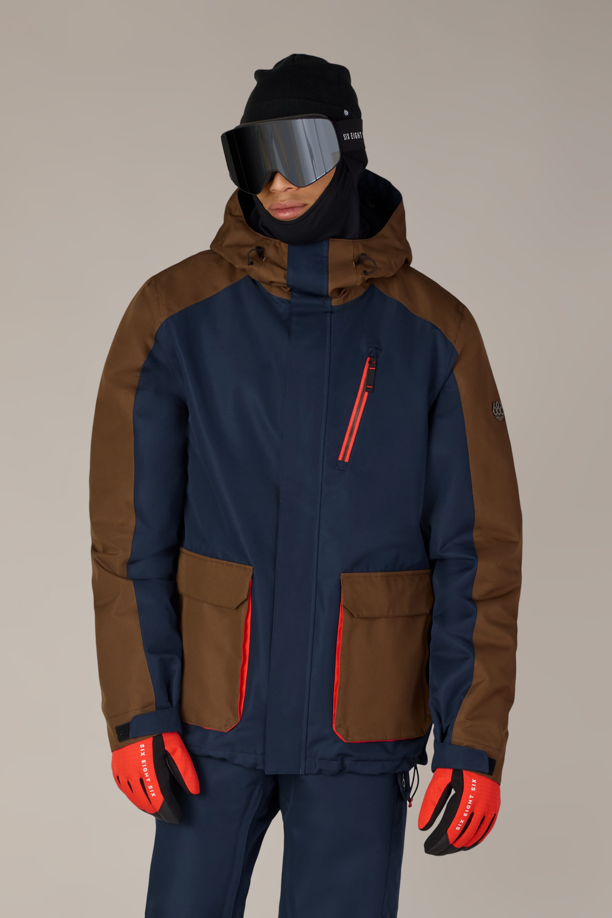 A person wearing the 686 Men’s Nightline™ Insulated Jacket in navy and brown with red accents, paired with matching pants, a black beanie, large ski goggles, and red gloves, stands against a plain background.