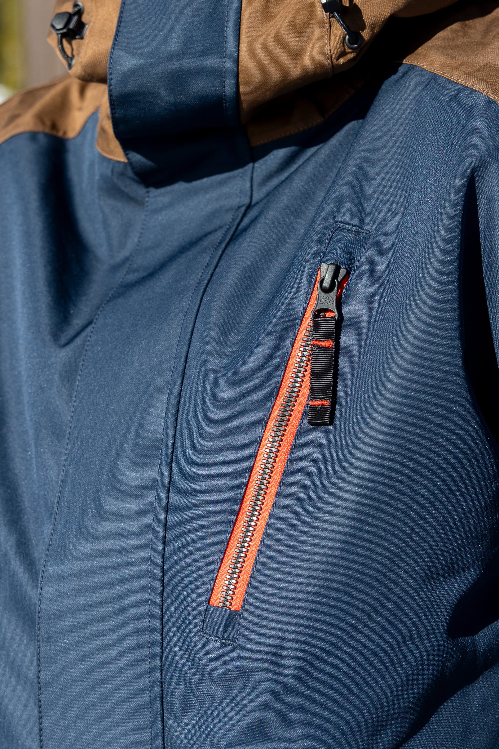 Here is a revised version using your product data: Close-up of the 686 Men’s Nightline™ Insulated Jacket by 686, featuring a blue exterior with a brown hood and a red zipper on the chest pocket. The durable material is designed to provide warmth and protection in outdoor or cold weather conditions.