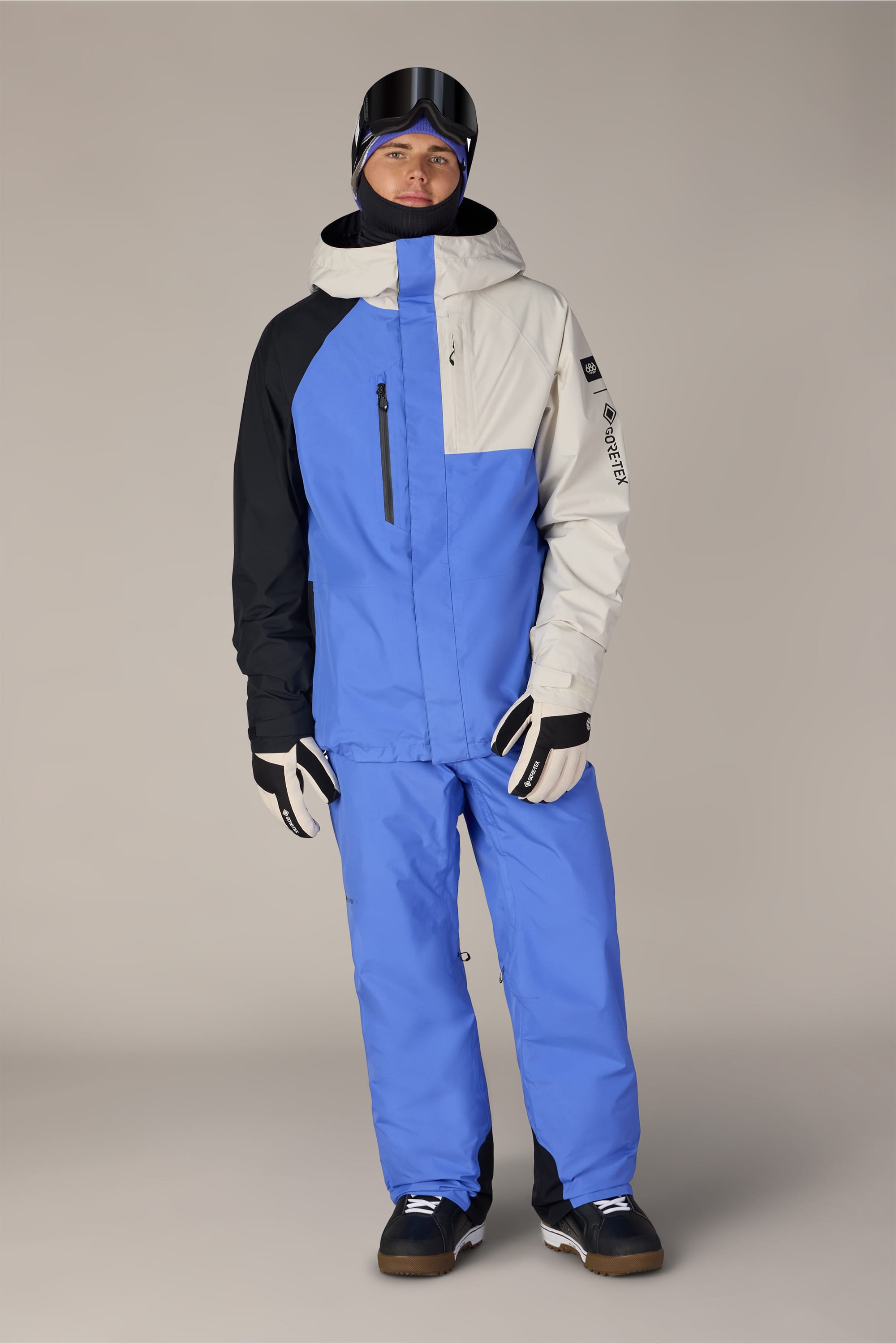 A person stands wearing a blue, black, and light gray ski jacket paired with 686 Men’s GORE-TEX® Core Shell Pant in blue by 686, along with black snow boots, white gloves, a black helmet, and ski goggles. The background is plain and neutral.