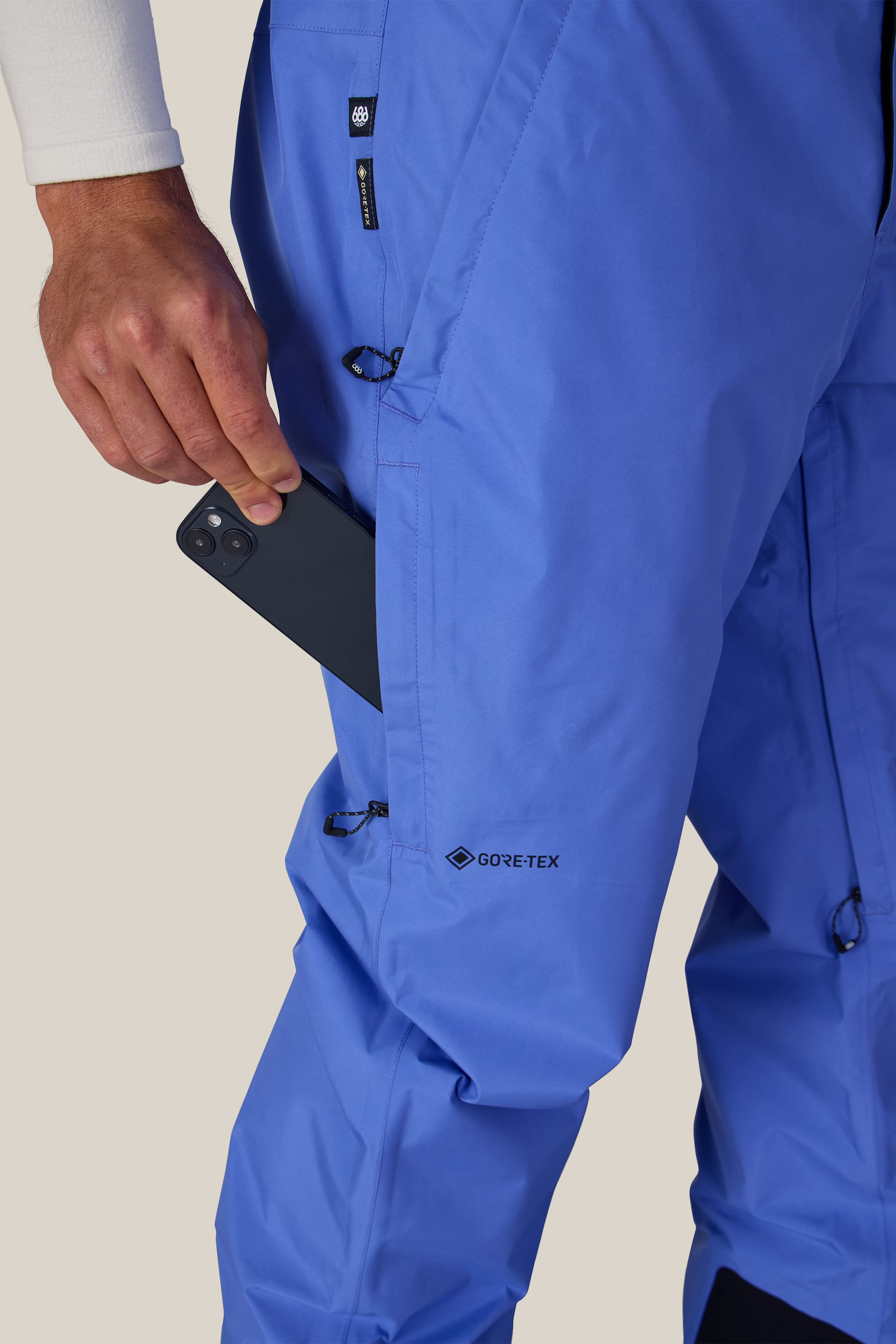 Wearing the 686 Men’s GORE-TEX® Core Shell Pant in bright blue, a person slips a black smartphone into the zippered thigh pocket; only their hand, part of their arm, and legs are visible against a neutral background.