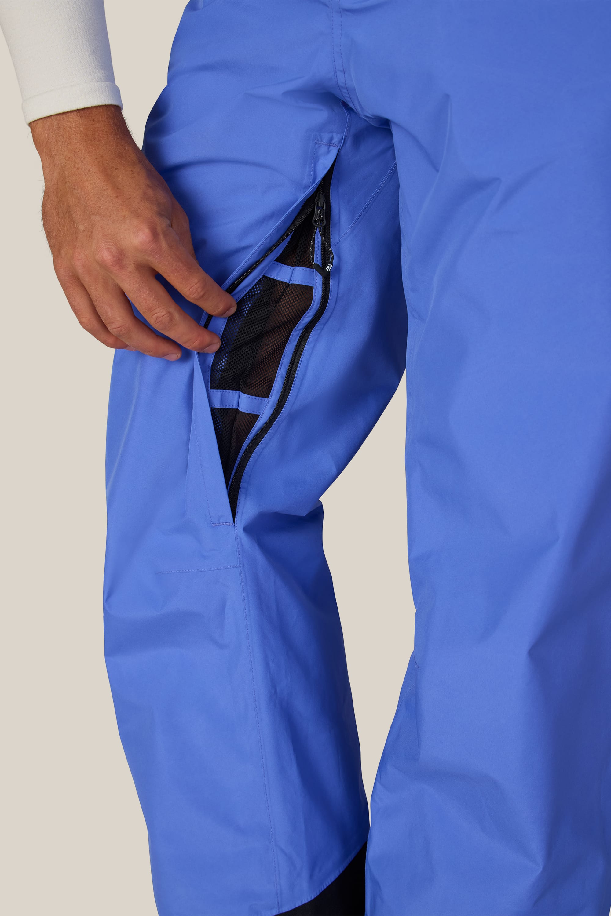 A person wearing the 686 Men’s GORE-TEX® Core Shell Pant unzips a mesh-vented side pocket, revealing part of their hand and white sleeve against a neutral background.