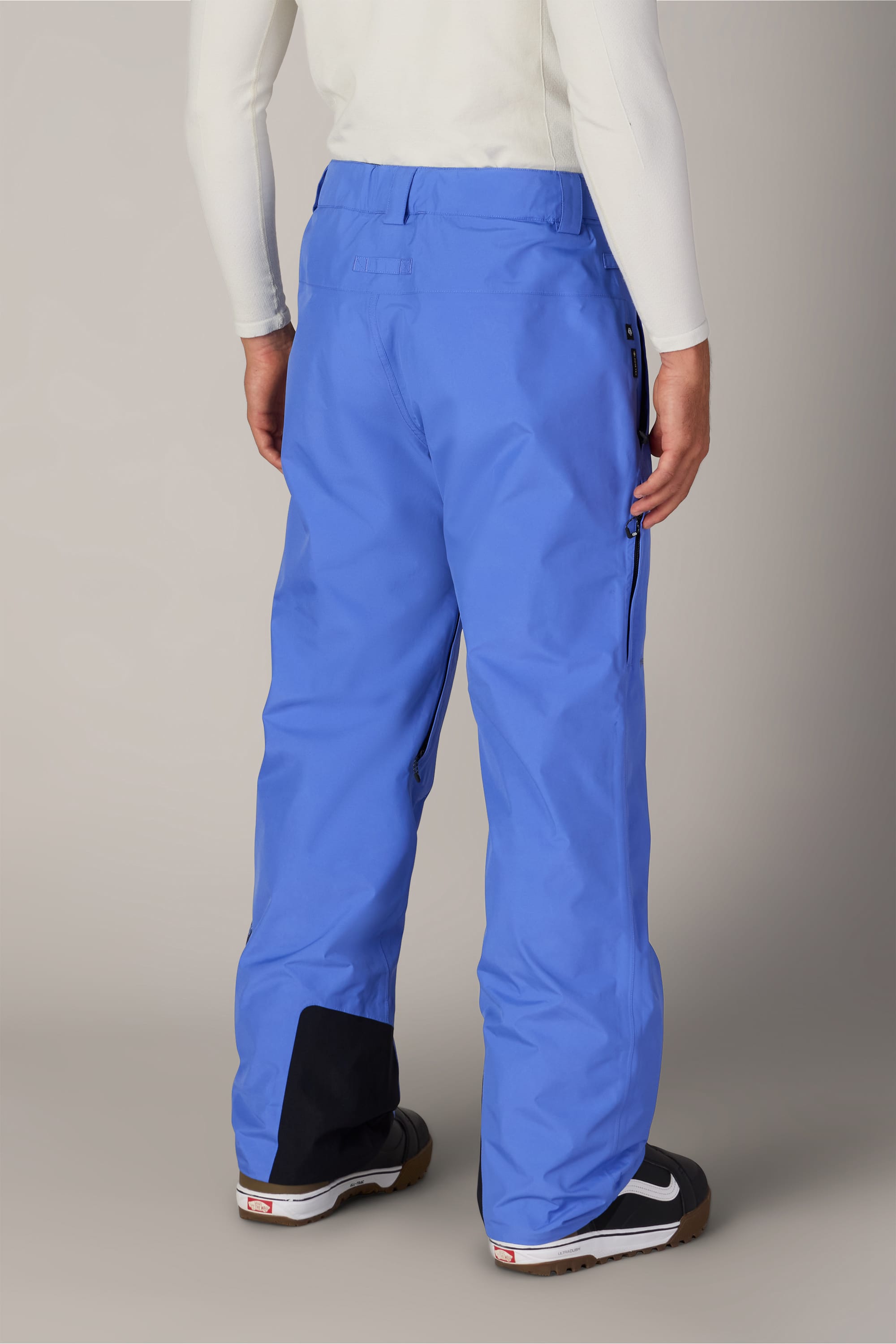 A person is shown from the back wearing the 686 Men’s GORE-TEX® Core Shell Pant in blue, featuring black patches on the lower legs. They pair it with a long-sleeve white top and black and white sneakers.