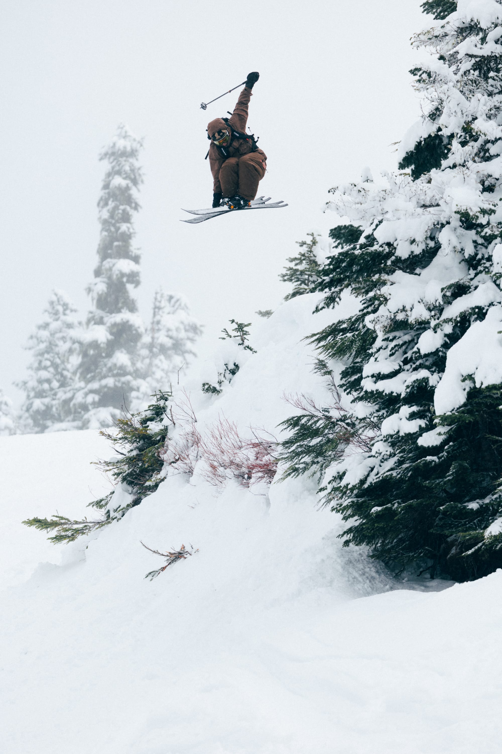 Wearing the 686 Men’s GORE-TEX® Core Shell Pant, a skier launches off a snowy ledge near snow-covered trees, captured mid-air with one arm raised on an overcast winter day.