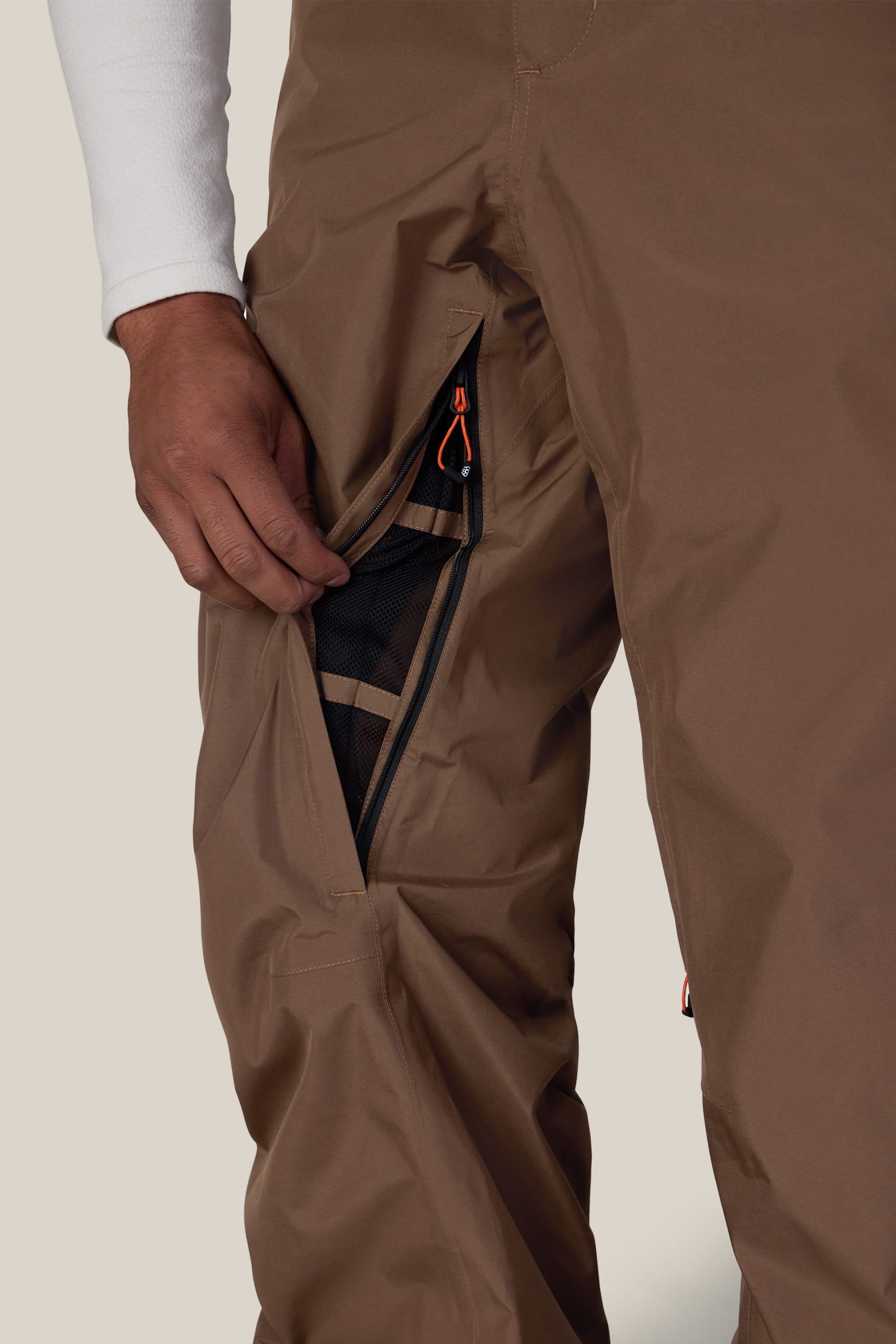 A person wearing the 686 Men’s GORE-TEX® Core Shell Pant in brown stands with a zippered vent open, revealing its black mesh lining. Their hand is holding the zipper, and they are dressed in a white long-sleeve shirt.