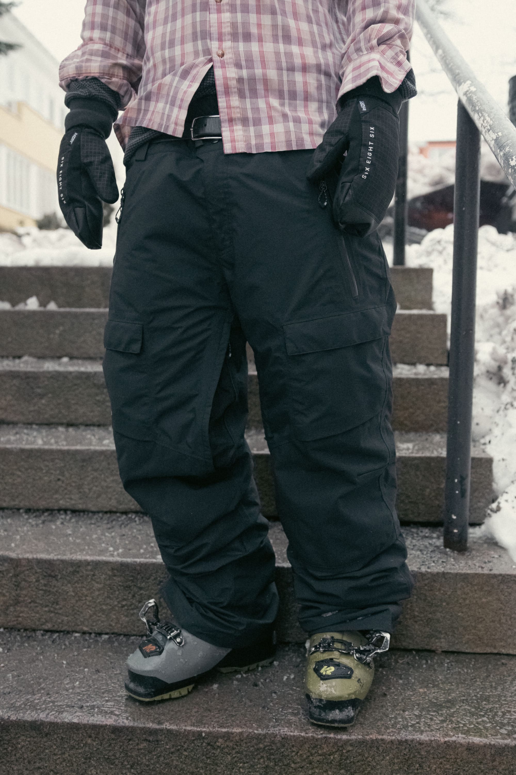 A person wearing the 686 Men’s GORE-TEX® Dispatch™ Shell Pant in black, mismatched ski boots (one gray, one green), black gloves, and a plaid shirt stands on outdoor stone steps surrounded by snow.
