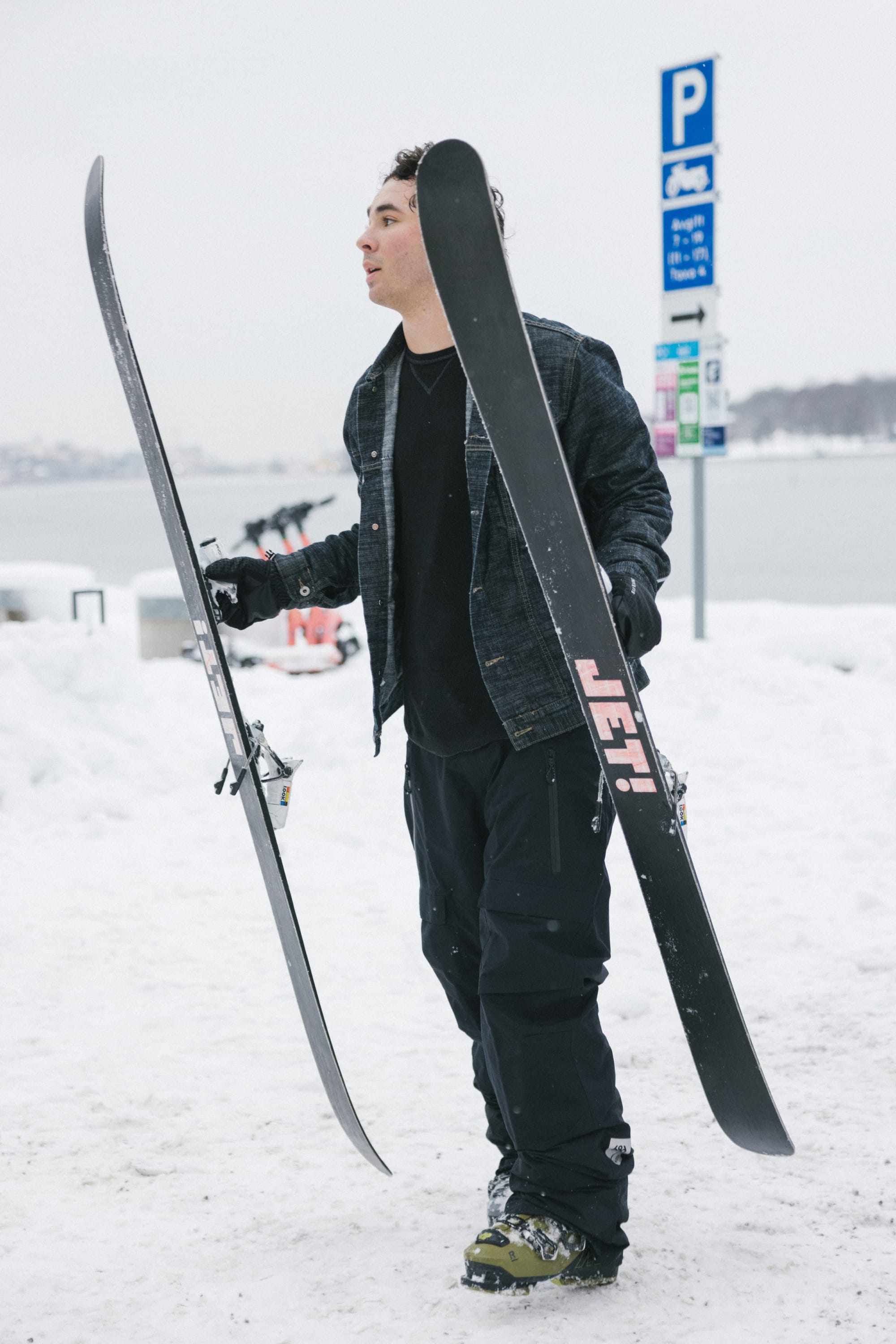 Wearing the 686 Men’s GORE-TEX® Dispatch™ Shell Pant, a person stands on snow holding two skis, with parking signs and a snowy landscape visible in the background.