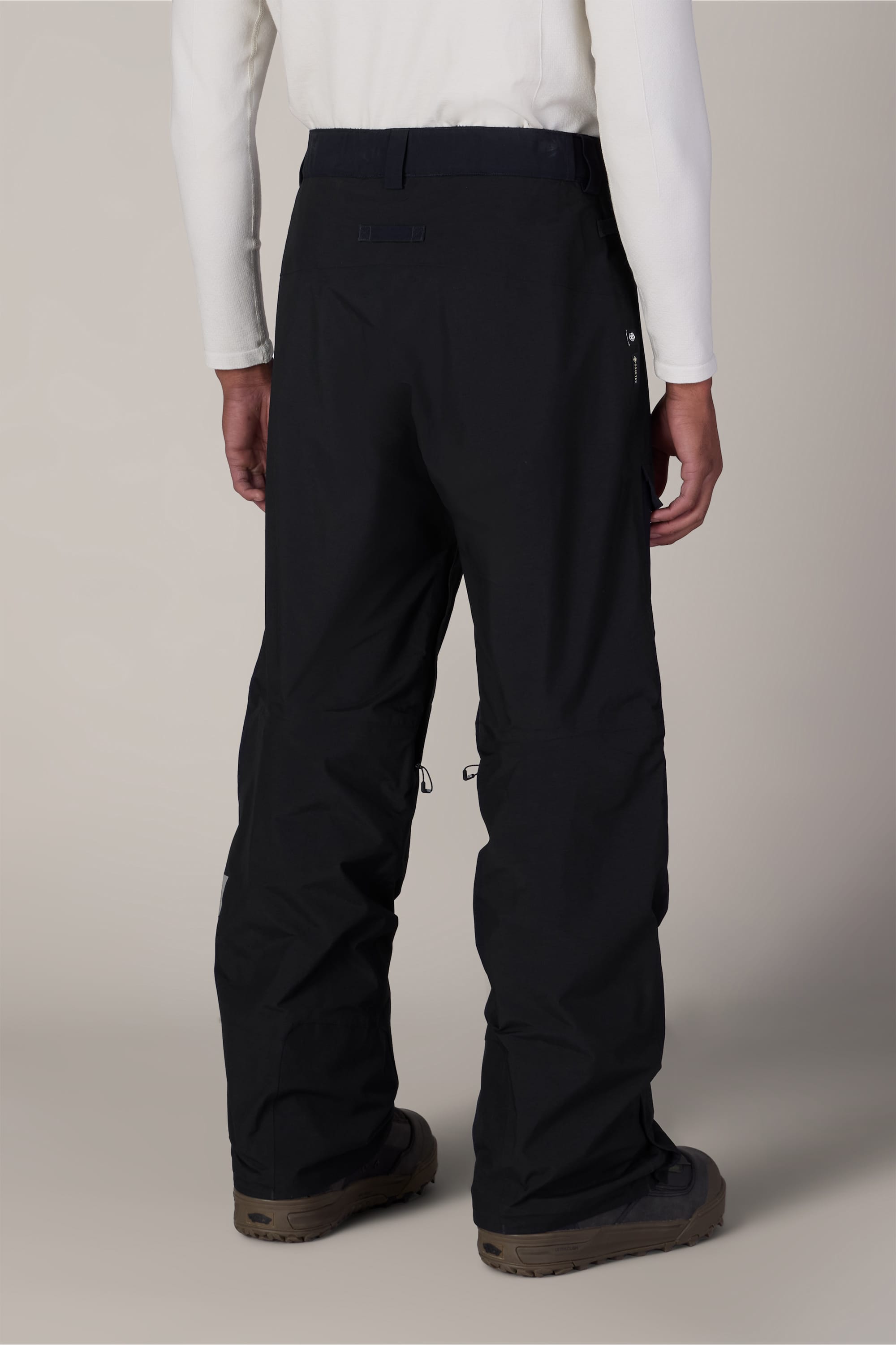 A person wearing a white long-sleeve shirt, brown shoes, and the 686 Men’s GORE-TEX® Dispatch™ Shell Pant in black is shown from the back, standing against a plain, light-colored background.