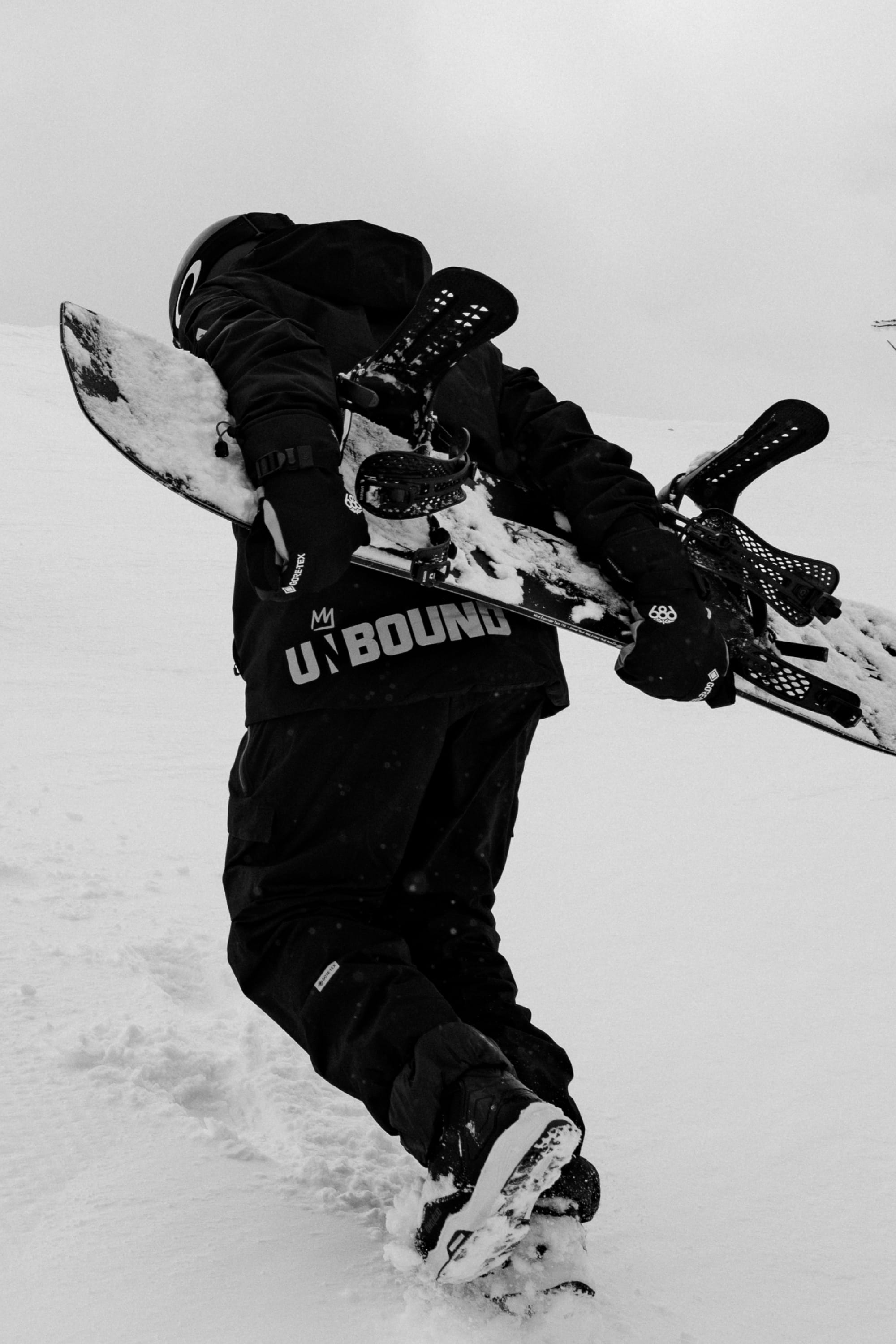 Wearing the 686 Men’s GORE-TEX® Dispatch™ Shell Pant, a person in winter gear carries a snowboard uphill on a snowy slope, with the word UNBOUND visible on their jacket in a black-and-white scene.