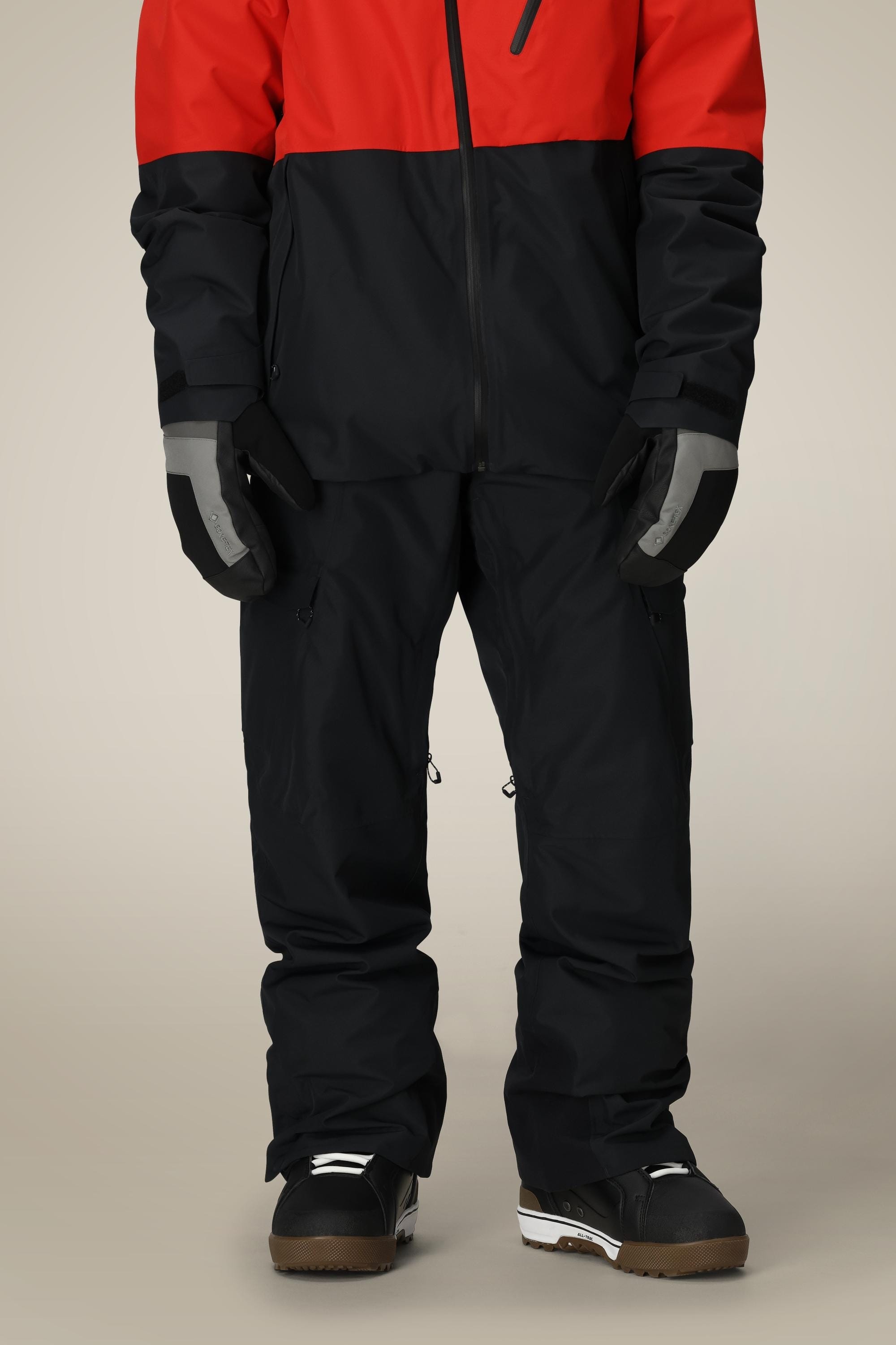 A person wearing a red and black jacket, 686 Mens Quantum Thermagraph® Pant by 686, gray and black gloves, and black and white snow boots stands against a plain background.