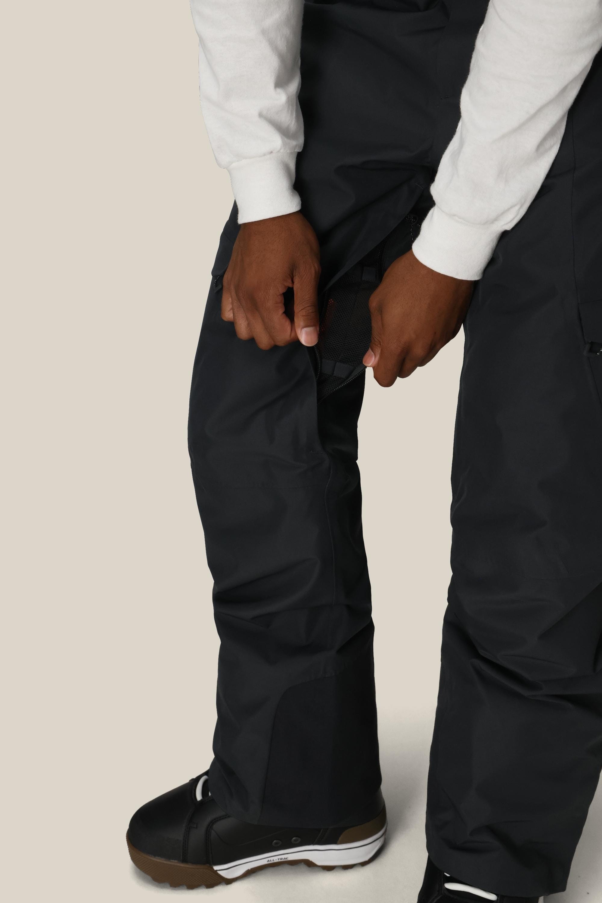 A person wearing 686 Mens Quantum Thermagraph® Pant in black with a white long-sleeve shirt and black snow boots adjusts a Velcro thigh strap. The background is plain and light-colored.