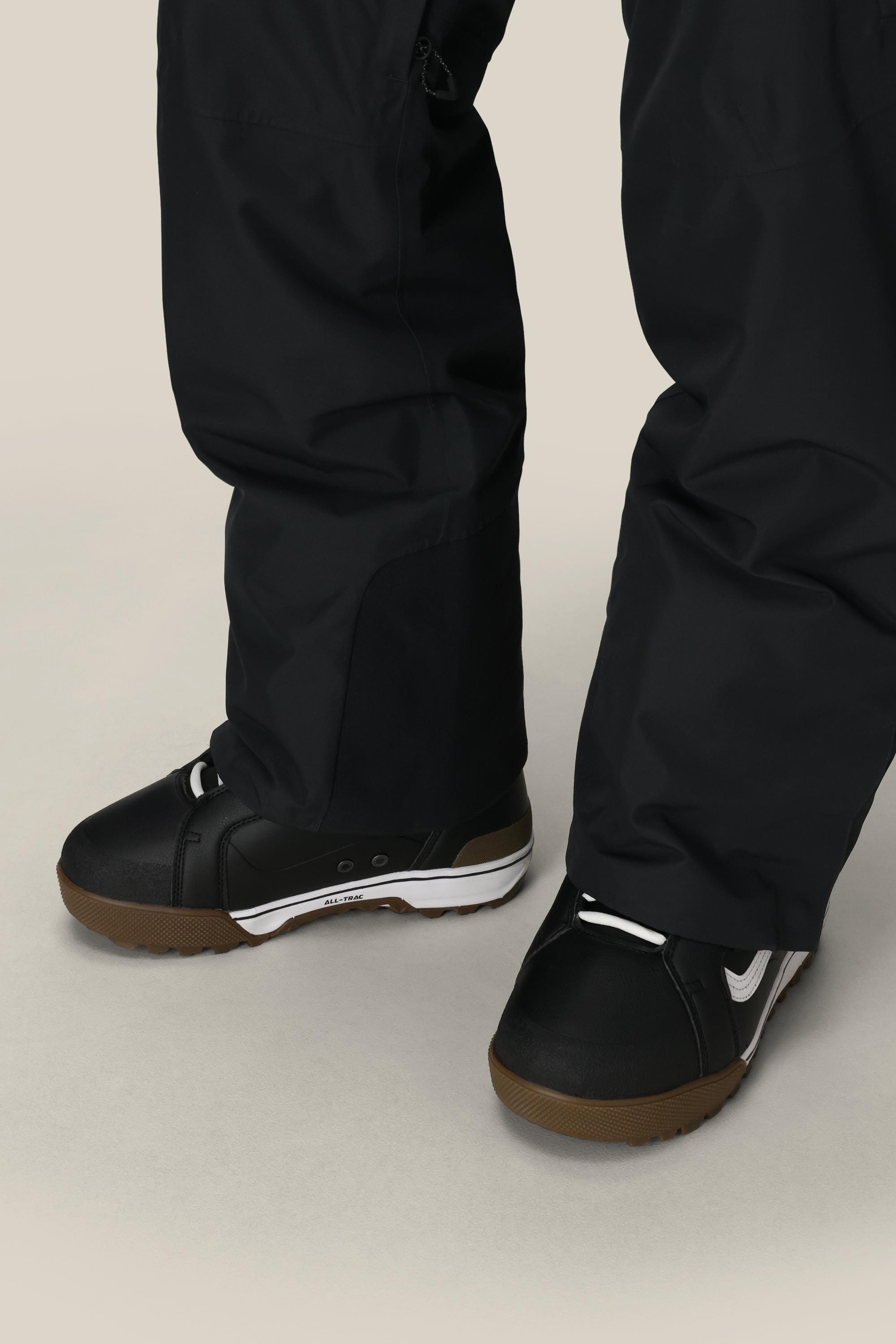 A close-up of a person wearing 686 Mens Quantum Thermagraph® Pant and black snow boots with white details and brown soles, standing on a light-colored surface.