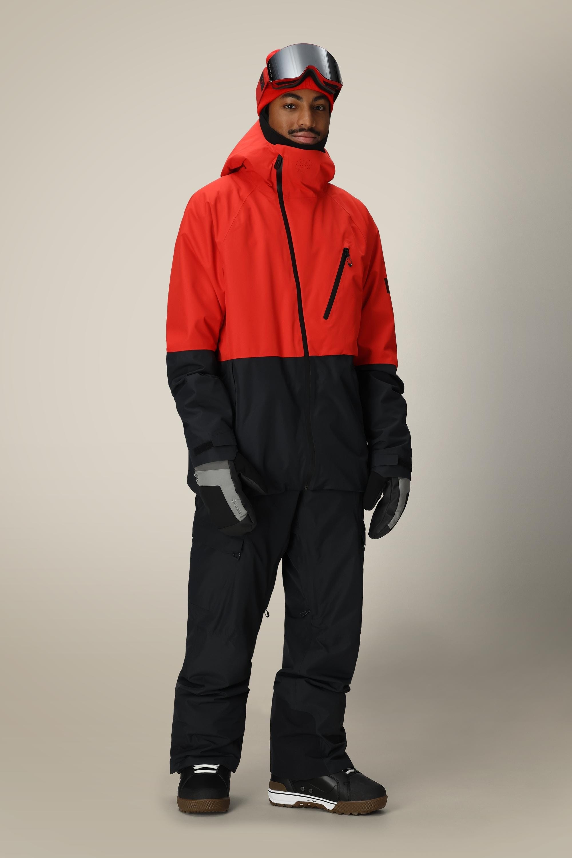 A person stands smiling slightly against a plain beige background, wearing a red and black ski jacket, 686 Mens Quantum Thermagraph® Pant by 686, gloves, ski boots, and red ski goggles on their head.