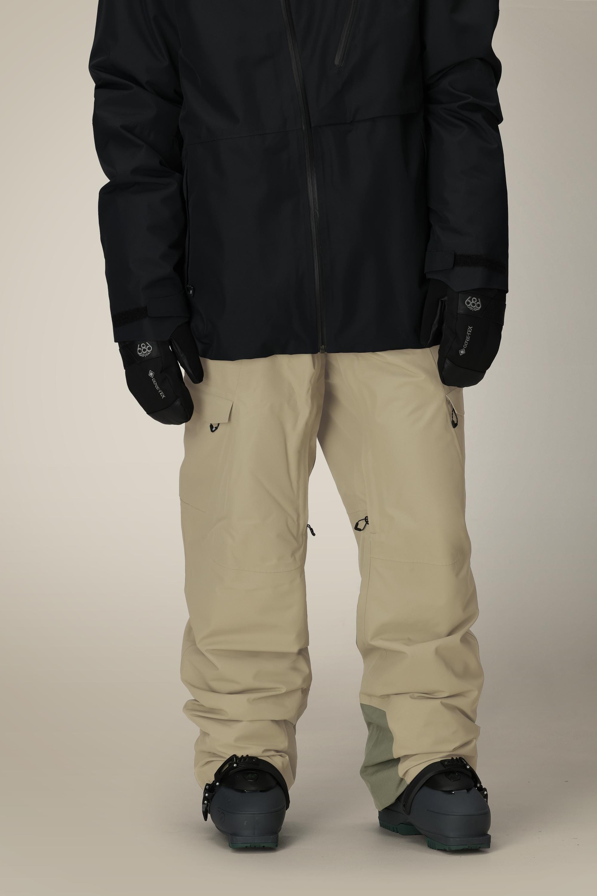 A person stands against a plain background wearing black gloves, a black jacket, black boots, and 686 Mens Quantum Thermagraph® Pant by 686 in beige. Their head is not visible in the image.