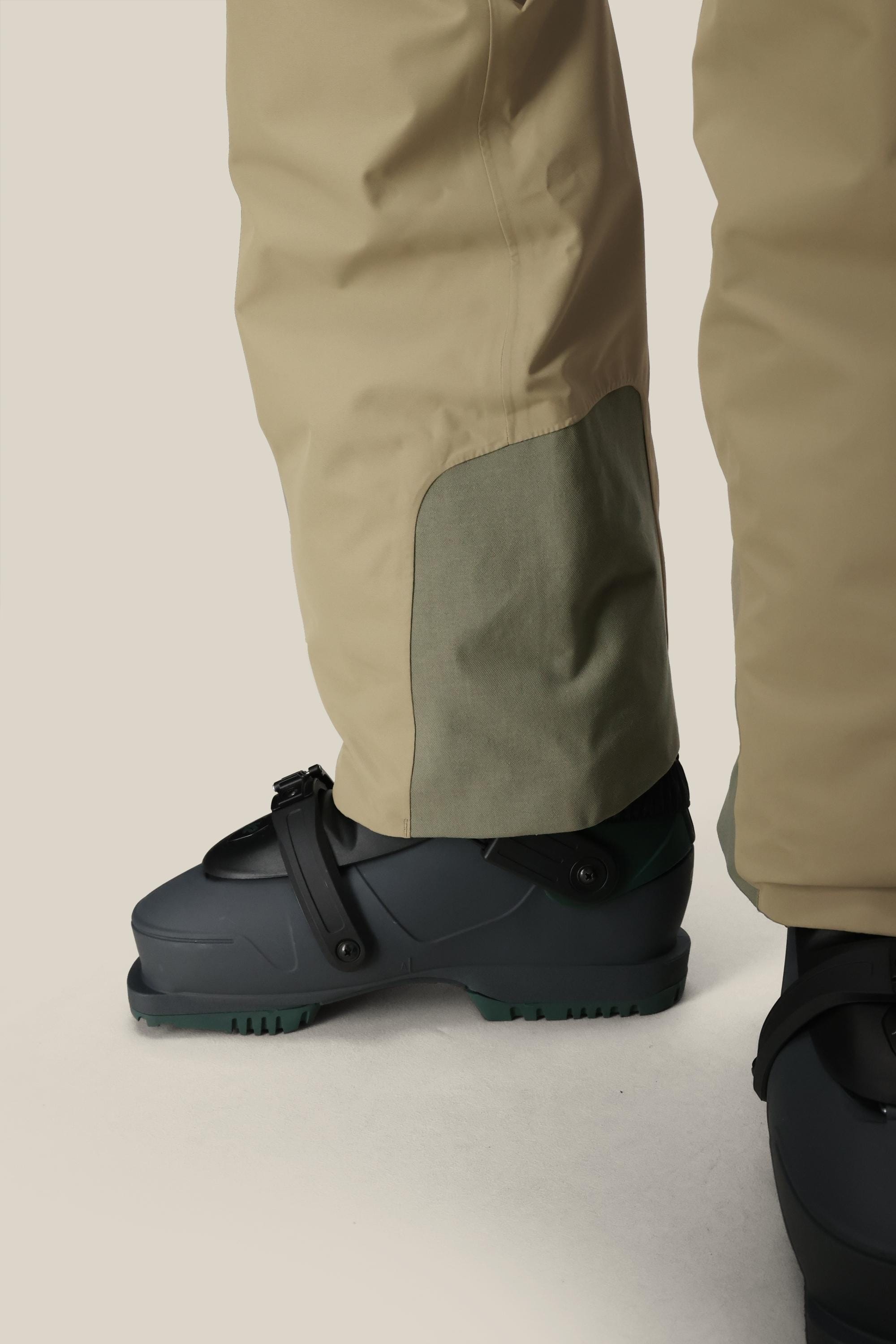 Close-up of a person wearing the 686 Mens Quantum Thermagraph® Pant in beige with a reinforced ankle patch, paired with black ski boots featuring green soles. The background is plain and light-colored. Brand: 686.