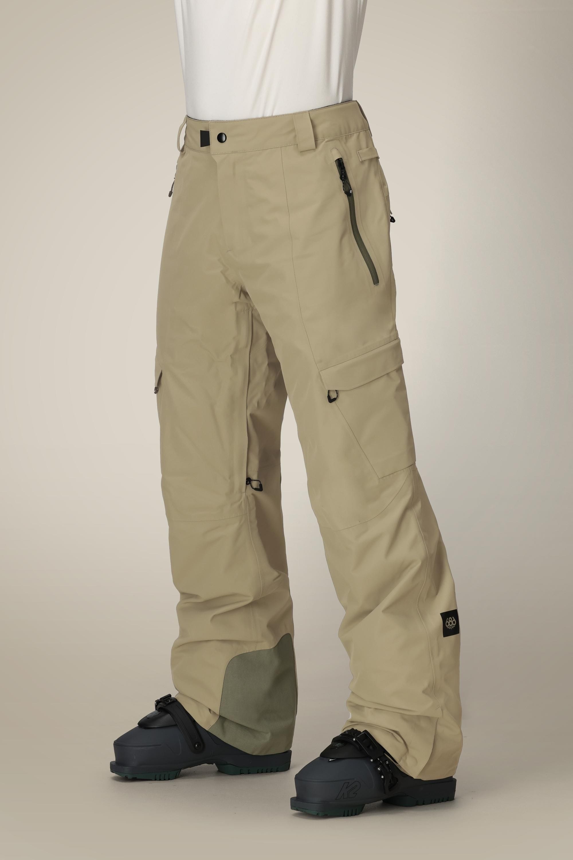 A person wearing 686 Mens Quantum Thermagraph® Pant in beige with black zippers and pockets, paired with a white shirt and black snowboard boots, stands against a plain light background.
