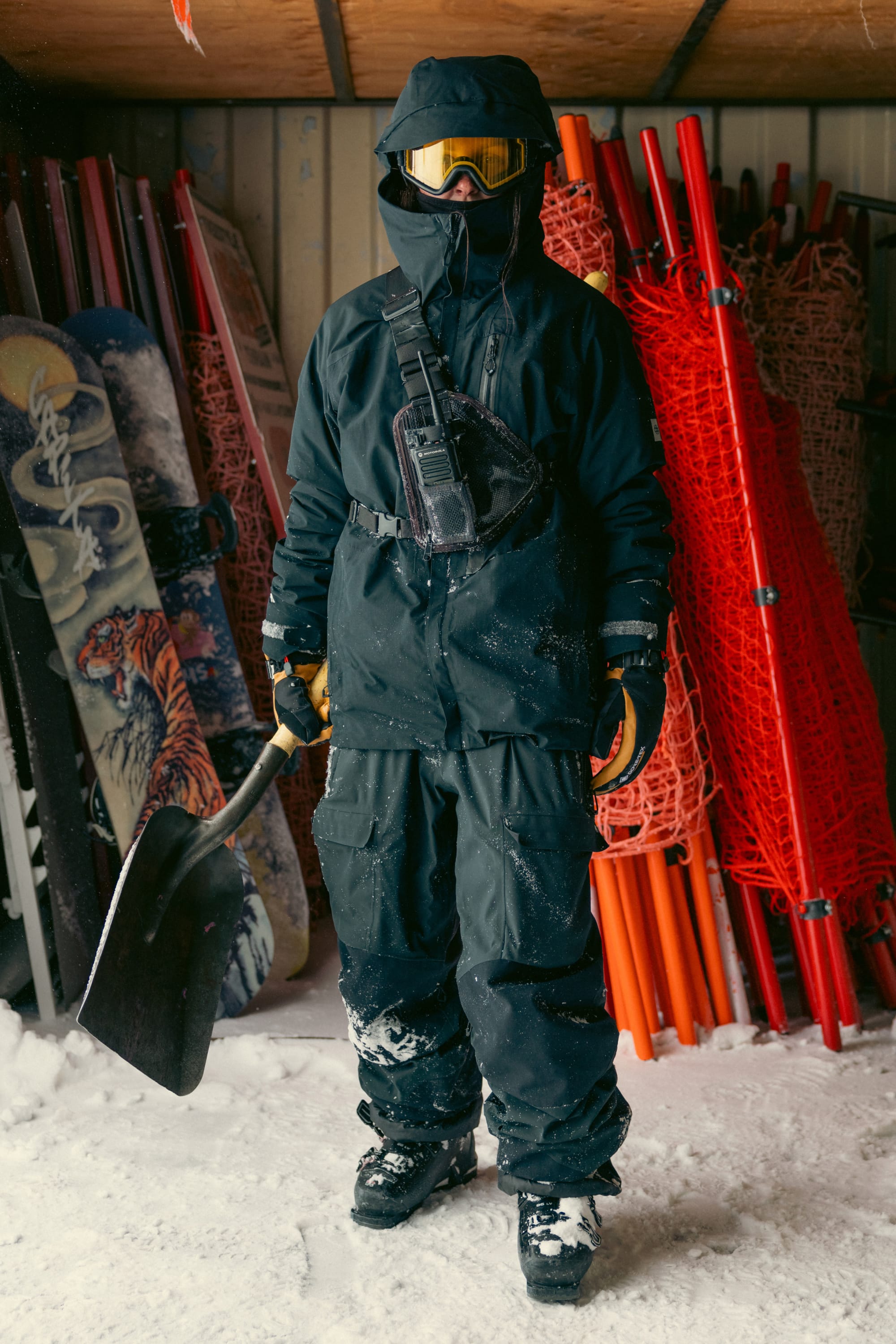Wearing the 686 Men’s GORE-TEX® Dispatch™ Shell Bib, a person in black snow gear, gloves, and goggles stands in a snowy shed holding a shovel, with snowboards and orange safety nets stacked in the background.