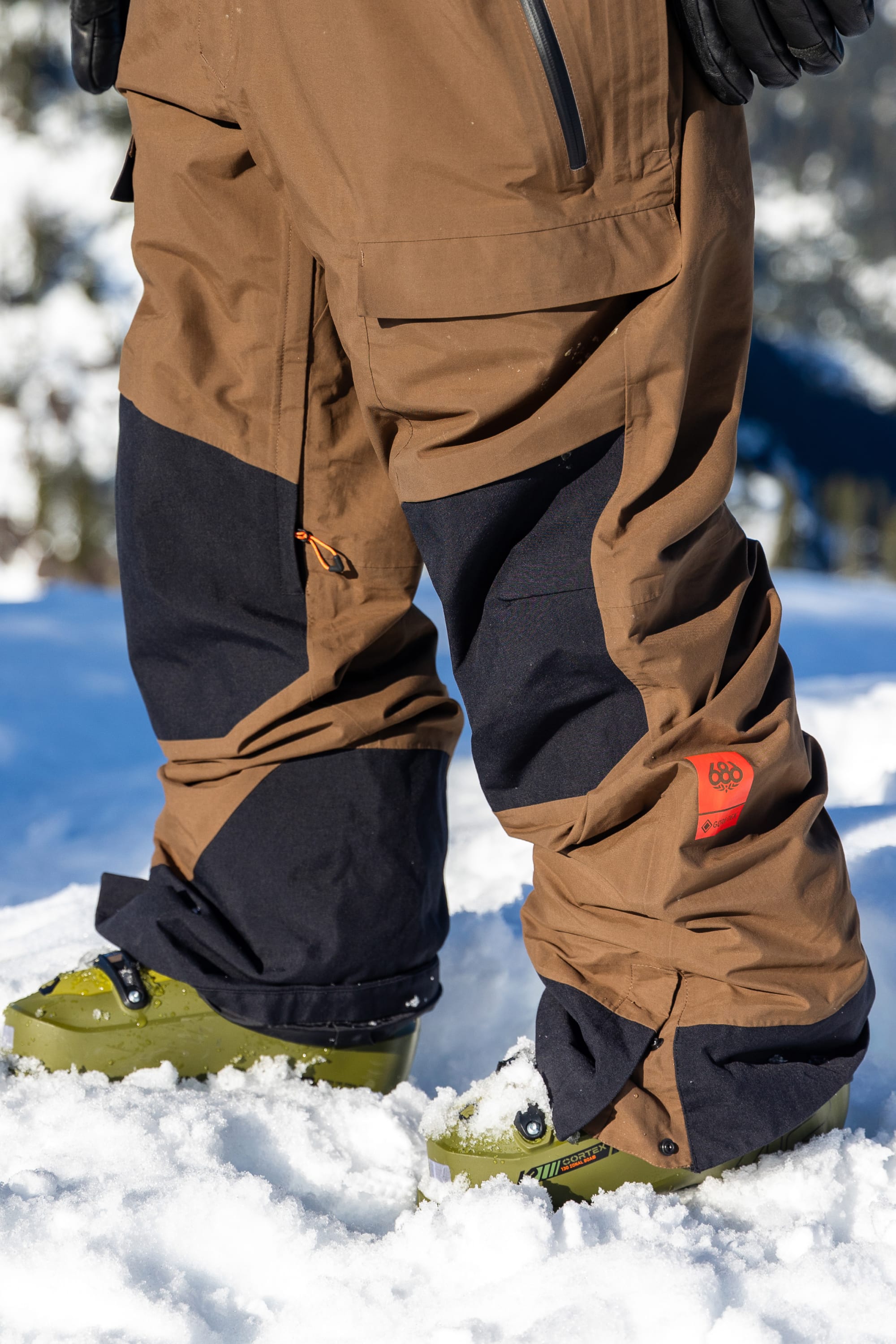 A person is standing in the snow wearing the 686 Men’s GORE-TEX® Dispatch™ Shell Bib by 686, along with green boots; gloves and a jacket are partially visible.