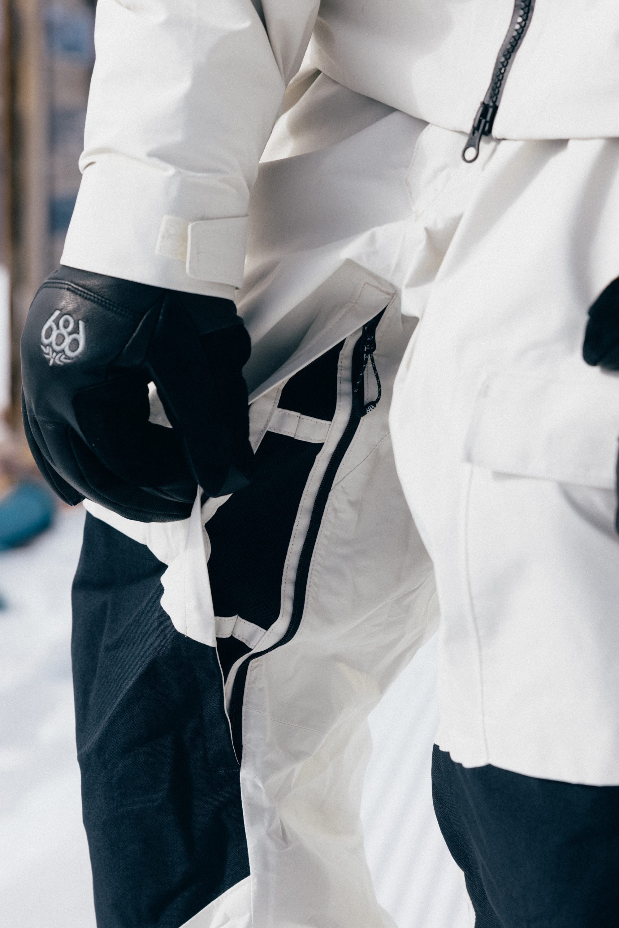 A person wearing black gloves and a white and black jacket unzips a side zipper on matching 686 Men’s GORE-TEX® Dispatch™ Shell Bib snow pants by 686, while standing in a snowy outdoor setting.