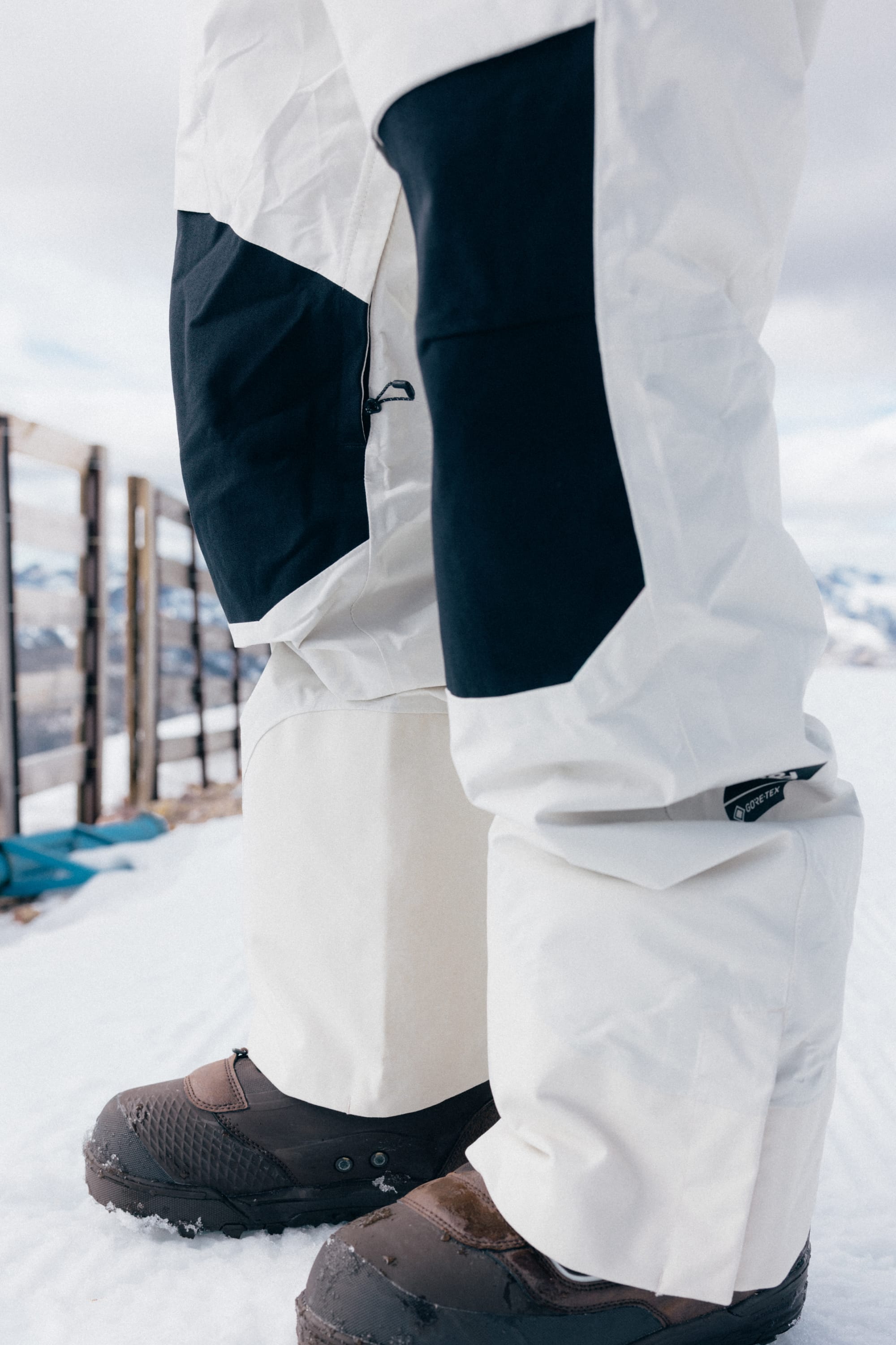 A close-up of a person wearing the 686 Men’s GORE-TEX® Dispatch™ Shell Bib snow pants and black snow boots standing on snow, with a wooden fence and snowy mountains in the background.