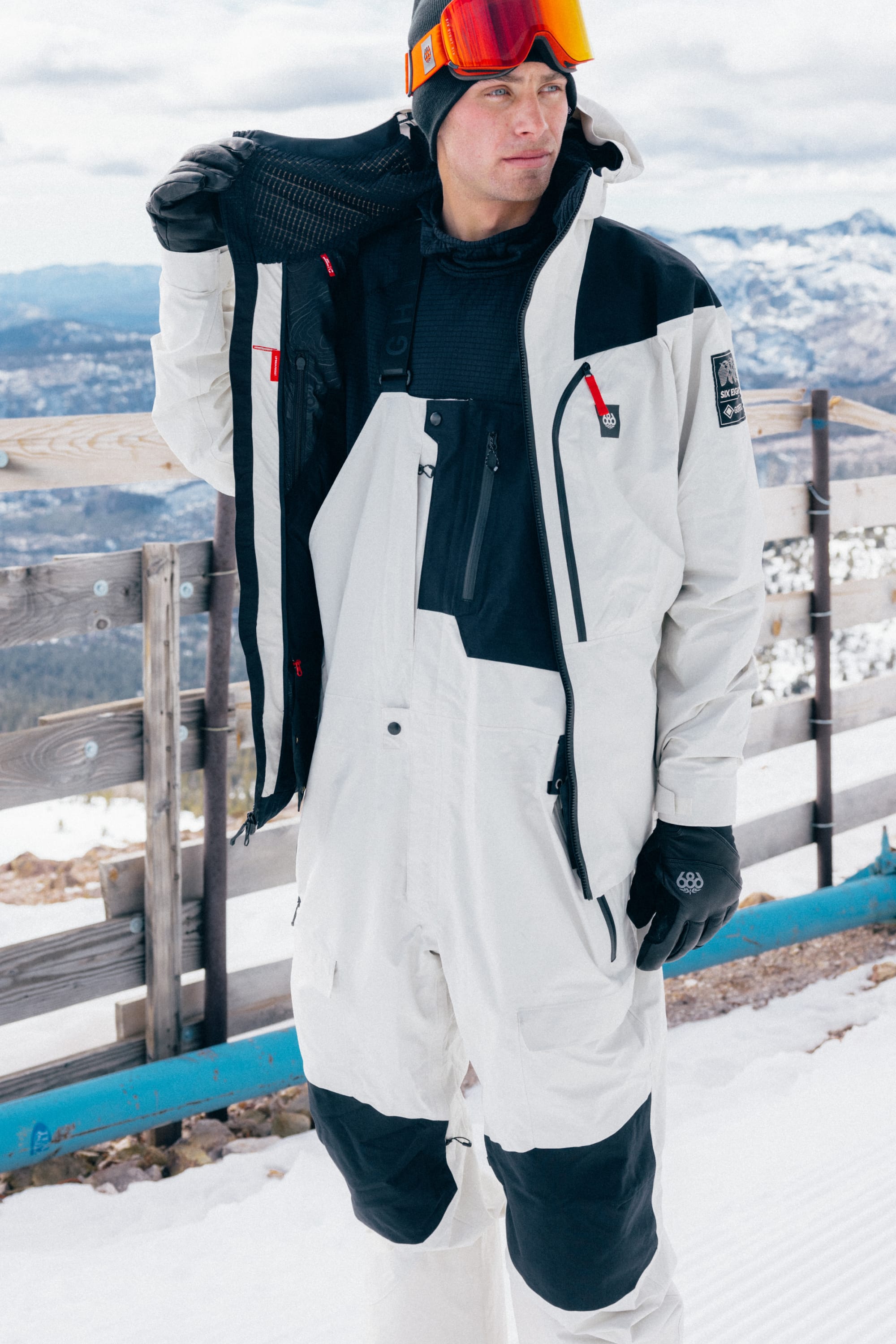 Wearing the 686 Men’s GORE-TEX® Dispatch™ Shell Bib, a person stands on snow near a wooden fence, orange goggles reflecting the snowy mountains in the background.