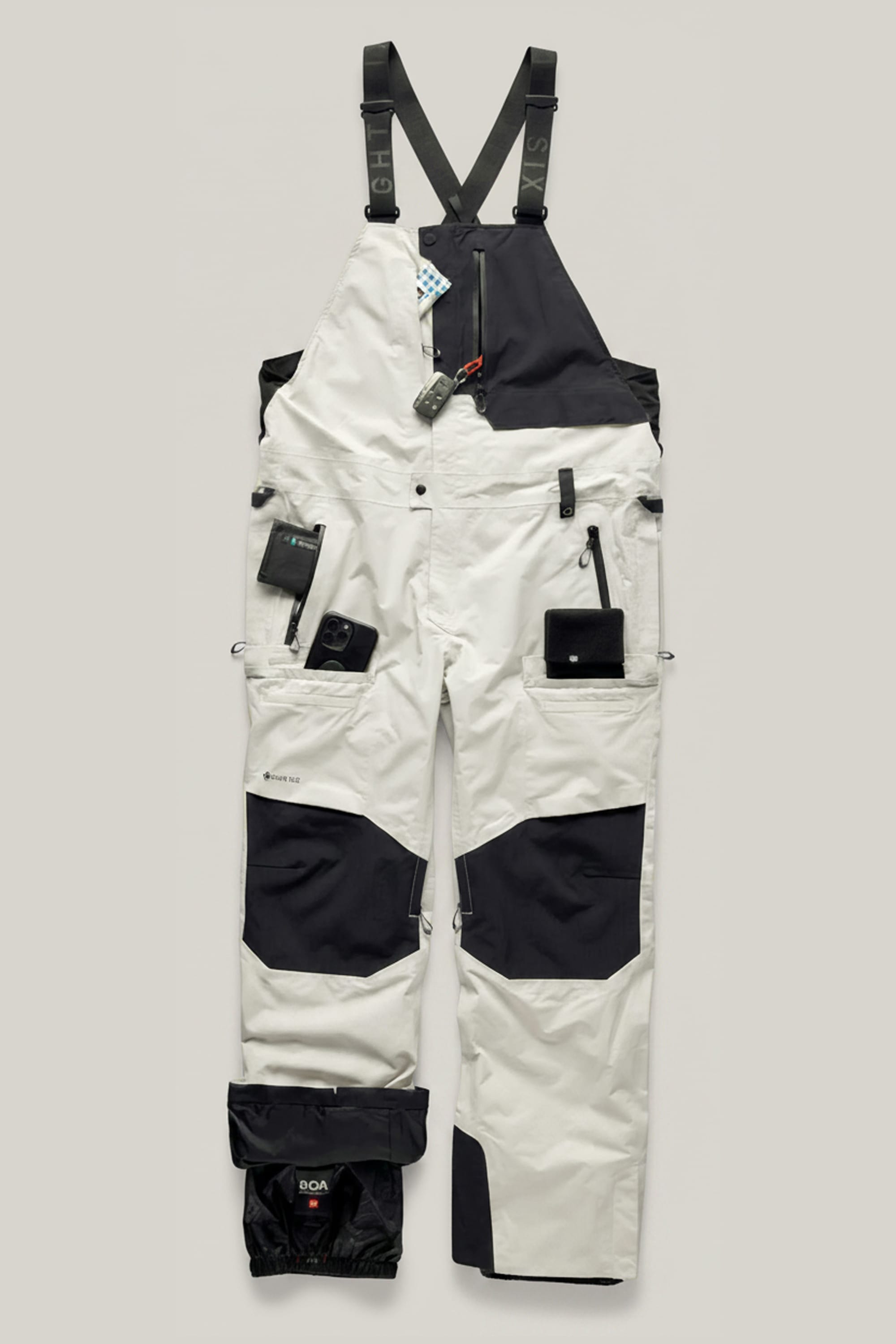 The 686 Men’s GORE-TEX® Dispatch™ Shell Bib by 686 is a black and white bib-style snow pant featuring multiple pockets, a chest clip, attached suspenders, and compartments showcasing handy gadgets like a multitool, camera, and wallet.