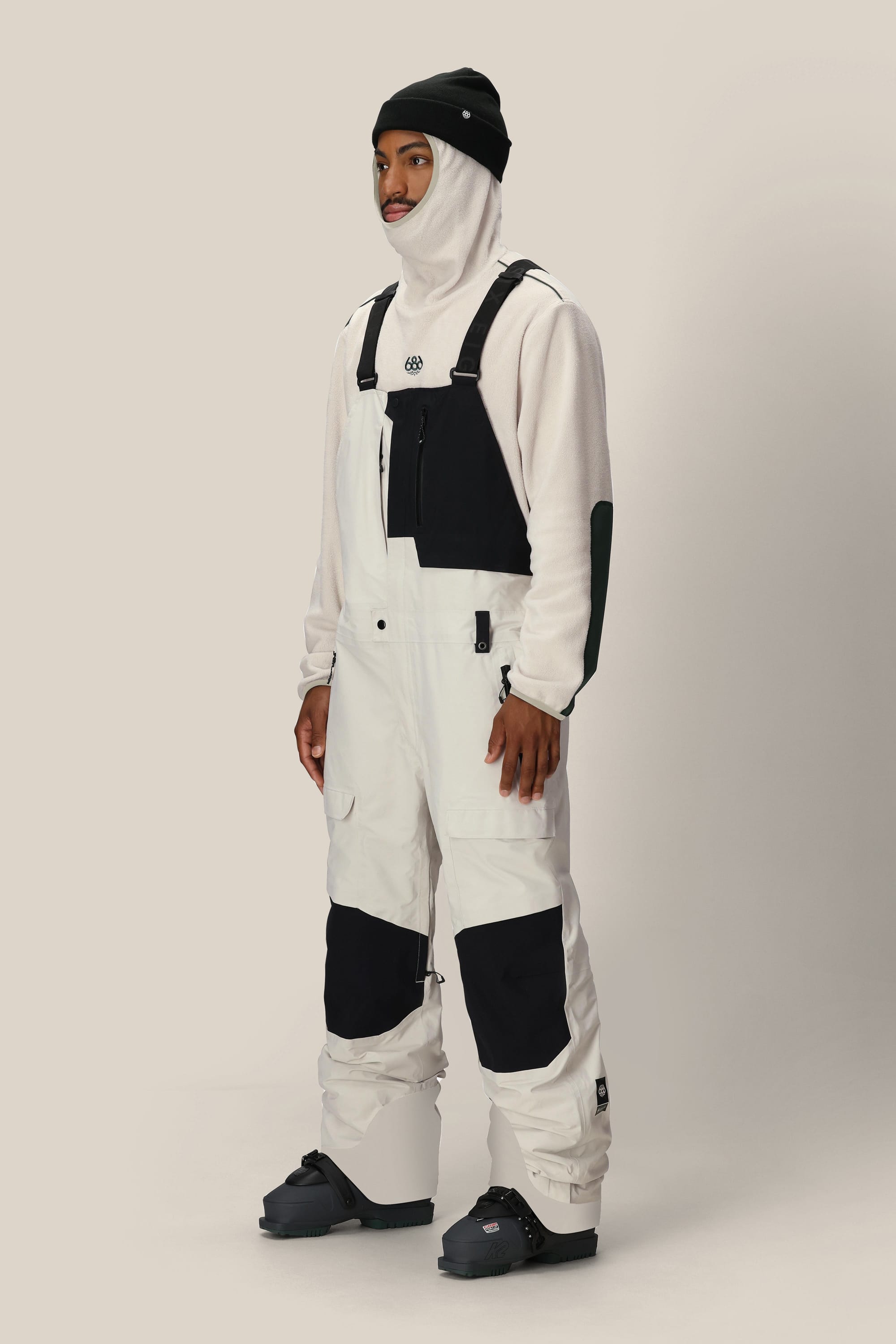 A person stands wearing a white hooded fleece and black beanie, paired with the 686 Men’s GORE-TEX® Dispatch™ Shell Bib by 686—white snow bib overalls featuring black knee patches. They complete the look with black snow boots and gaze to the side against a plain background.