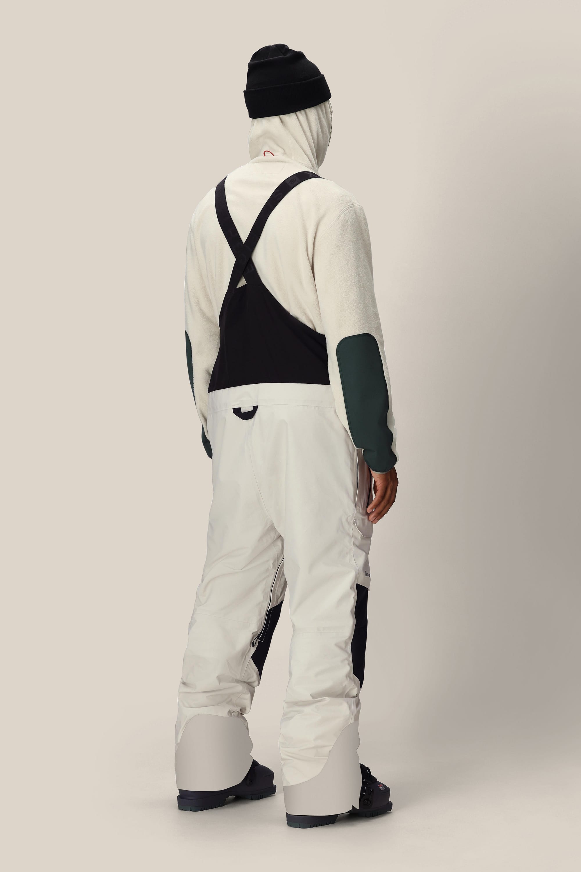 A person stands facing away, dressed in a white hoodie, black beanie, and the 686 Men’s GORE-TEX® Dispatch™ Shell Bib snow pants with black suspenders and patches from 686, paired with black snow boots, against a plain light background.