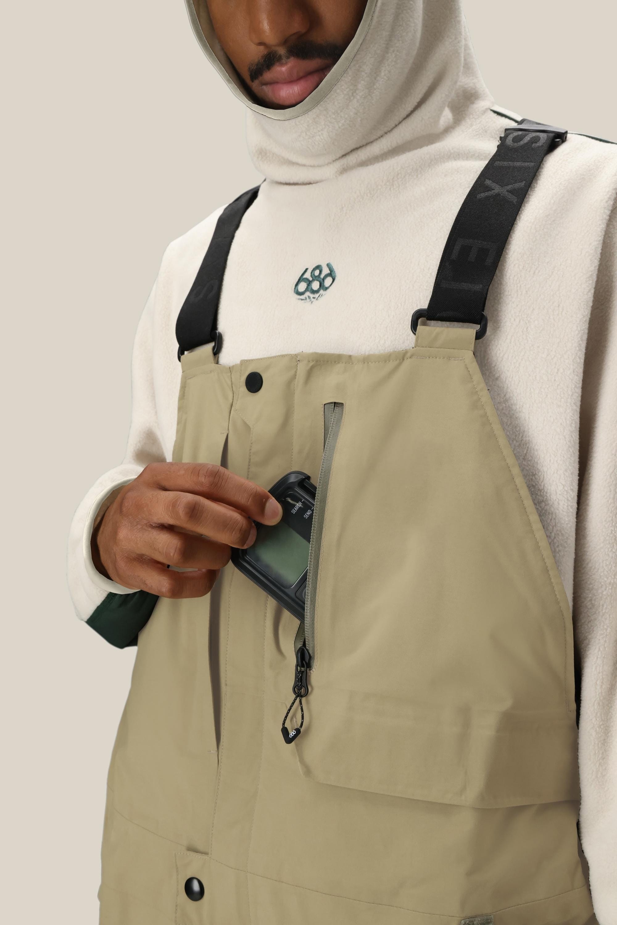 A person wearing the 686 Men's GORE-TEX Dispatch™ Shell Bib in light beige with black straps and a cream hoodie unzips a chest pocket to place a black phone inside. Only their lower face and upper body are visible.