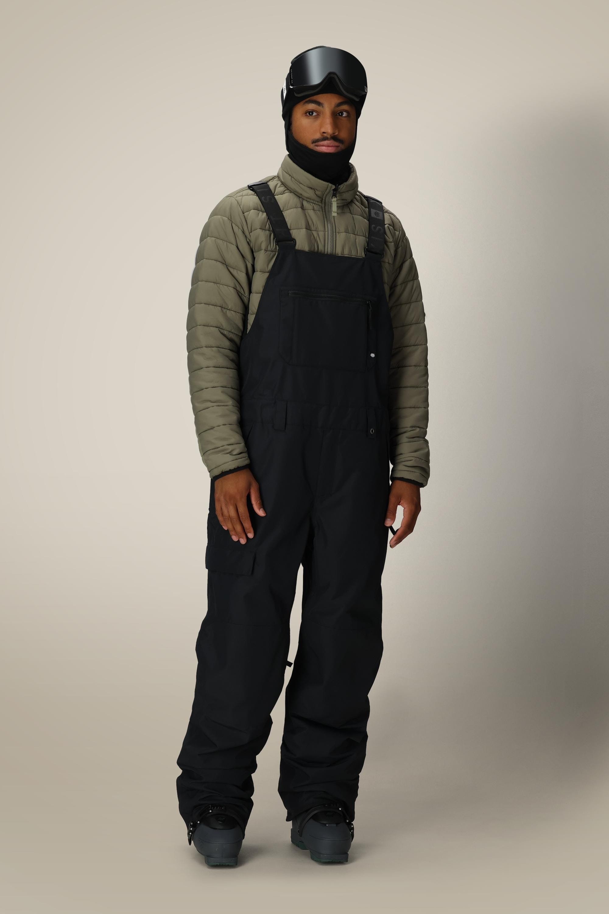A person stands facing forward against a plain light background, wearing a black ski helmet, the 686 Mens 20K Hot Lap Shell Bib by 686 made from waterproof woven polyester nylon spandex, a light olive green puffer jacket, and black snow boots.