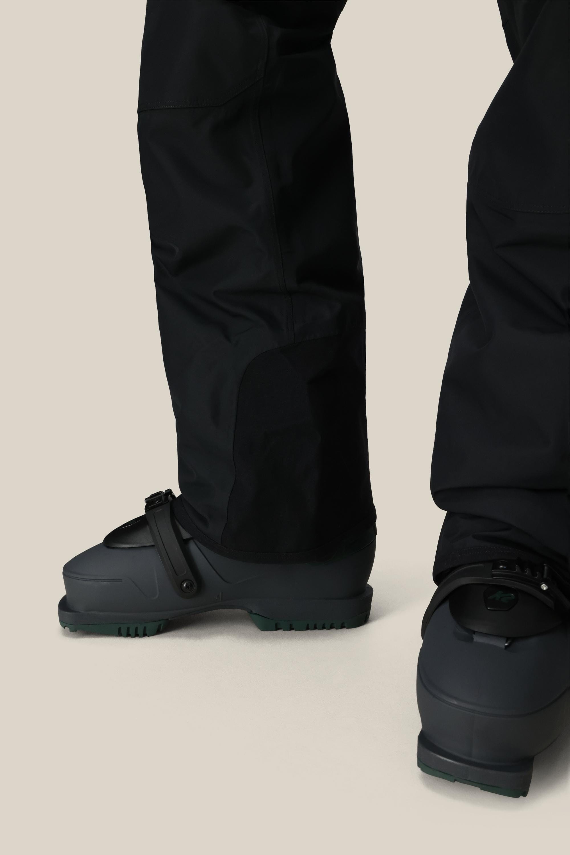 A close-up of two people wearing 686 Mens 20K Hot Lap Shell Bib snow pants by 686, paired with dark gray snow boots with bindings, standing on a light beige background.