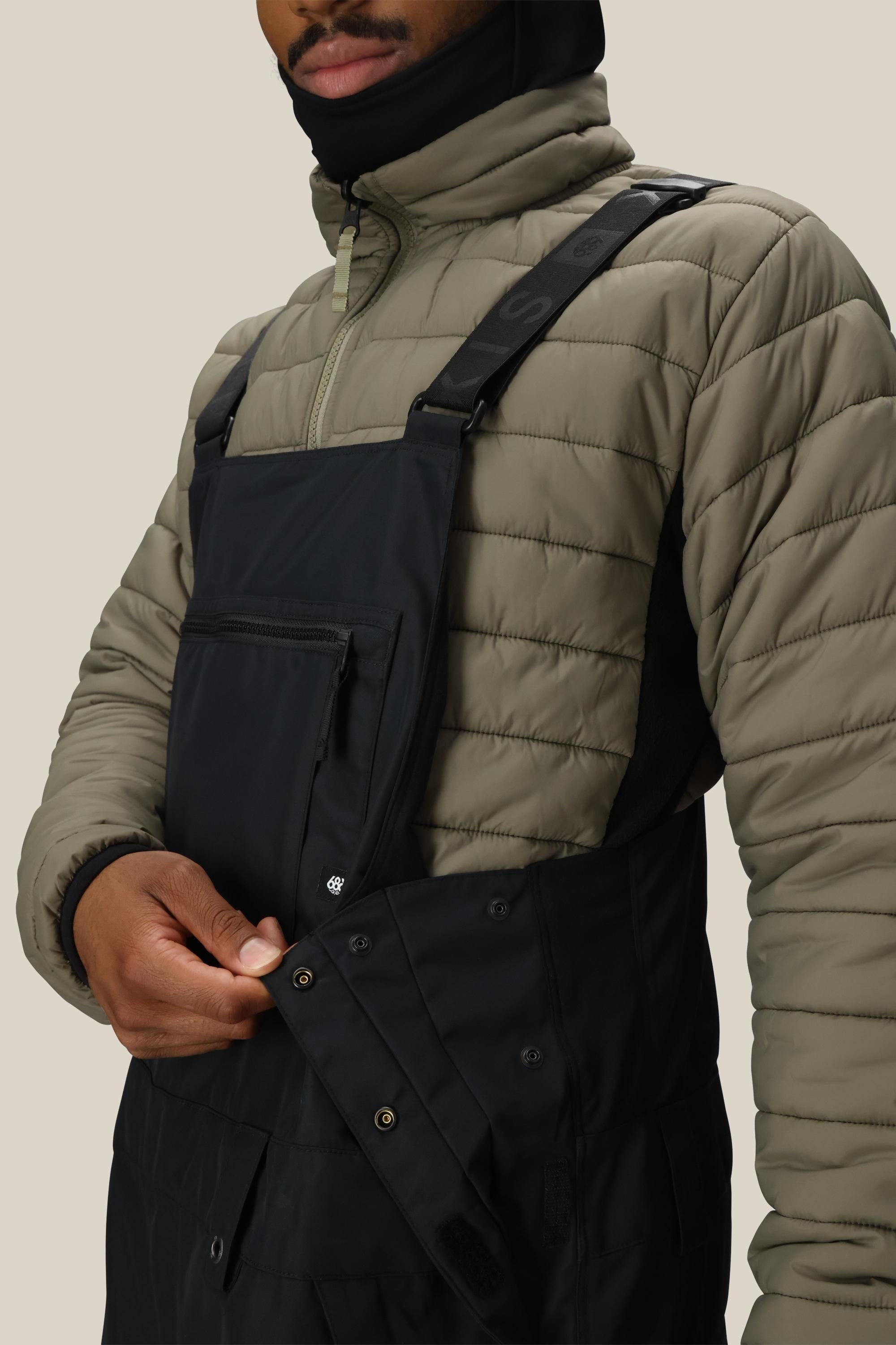 Wearing the 686 Mens 20K Hot Lap Shell Bib, a person in a khaki jacket demonstrates unbuttoning the side. The warm, waterproof bib from 686 looks perfect for outdoor activities, with part of the wearer’s face visible.