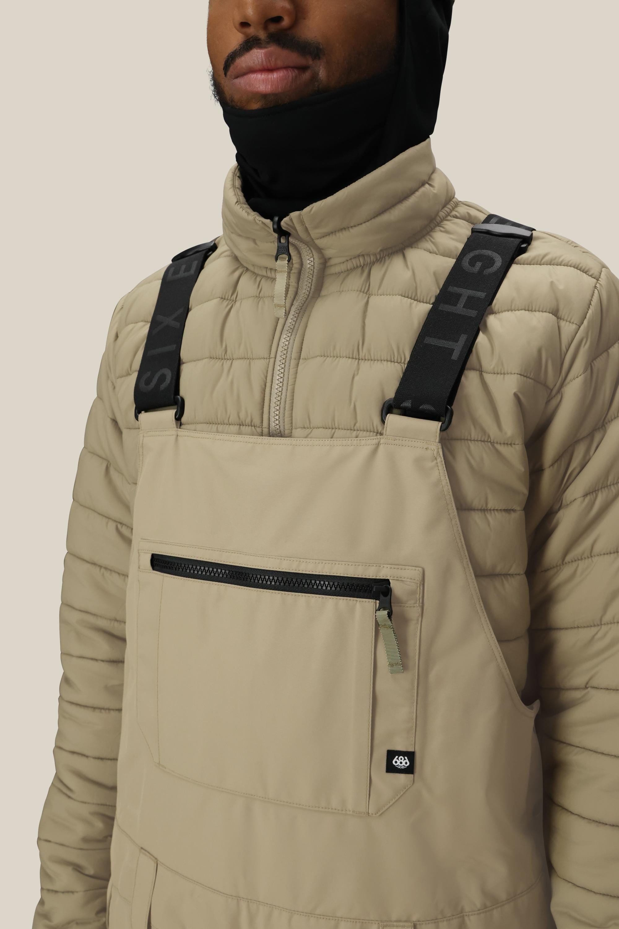 A person wears the 686 Mens 20K Hot Lap Shell Bib—beige waterproof overalls with a zip pocket, black SIX EIGHT straps—over a beige quilted jacket and pairs it with a black face covering.