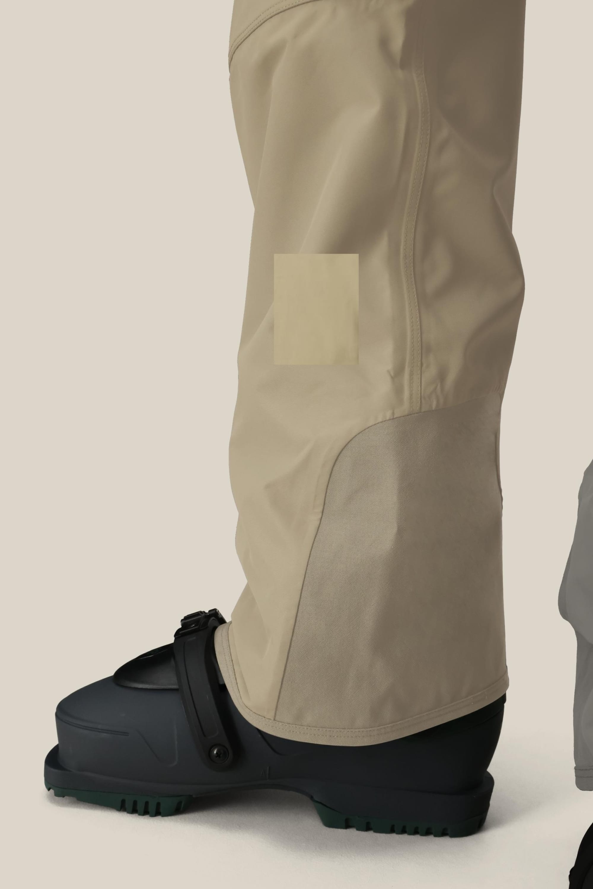 A close-up side view of a person wearing 686 Mens 20K Hot Lap Shell Bib pants and a black ski boot with green treads, set against a plain light background. Only the lower leg in the beige bib by 686 and boot are visible.